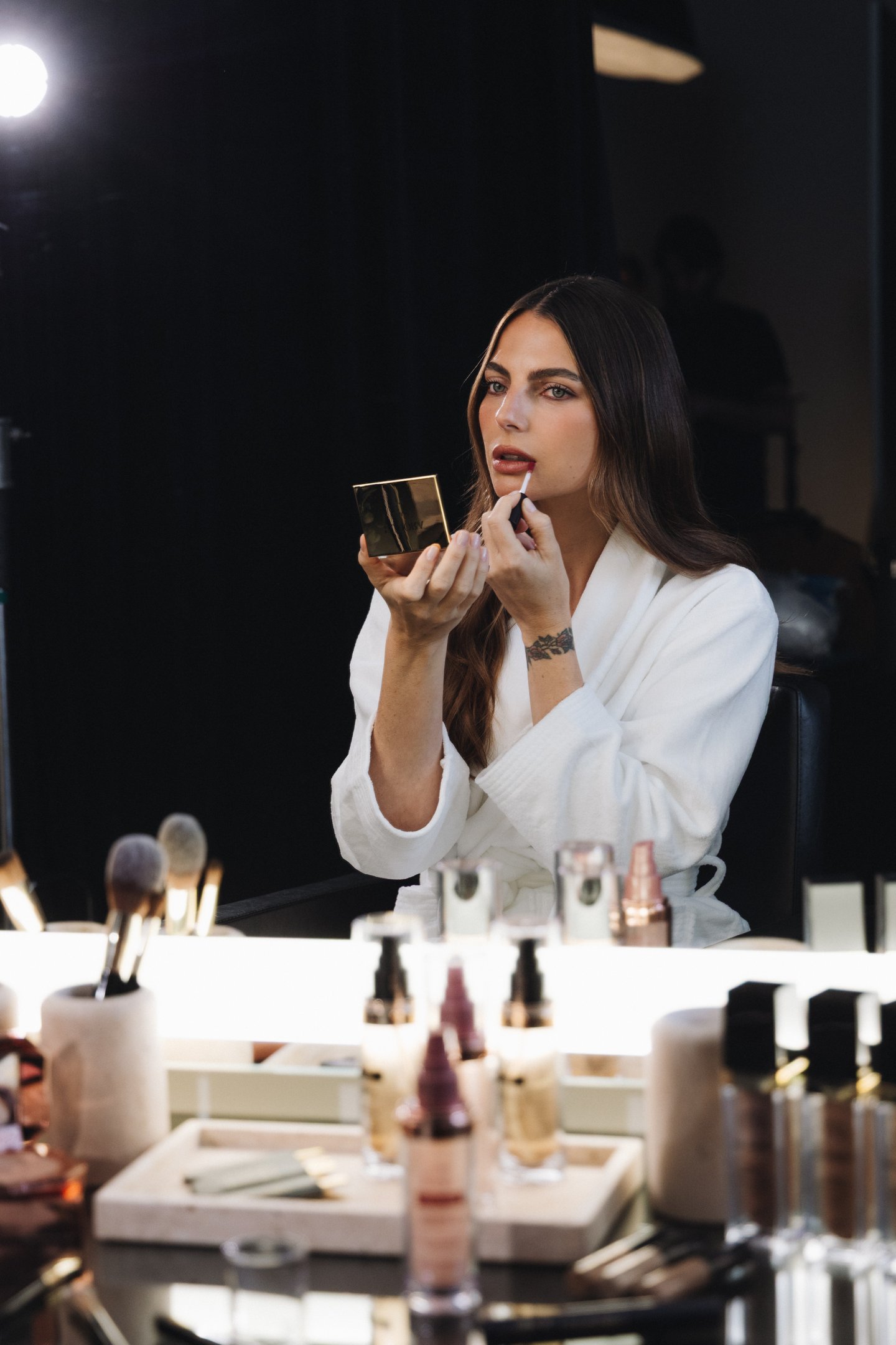 Kelly Piquet aplicando batom em um espelho, vestindo roupão branco. A mesa está cheia de produtos de beleza, como frascos e pincéis. WAG destaca a preparação para eventos de moda.