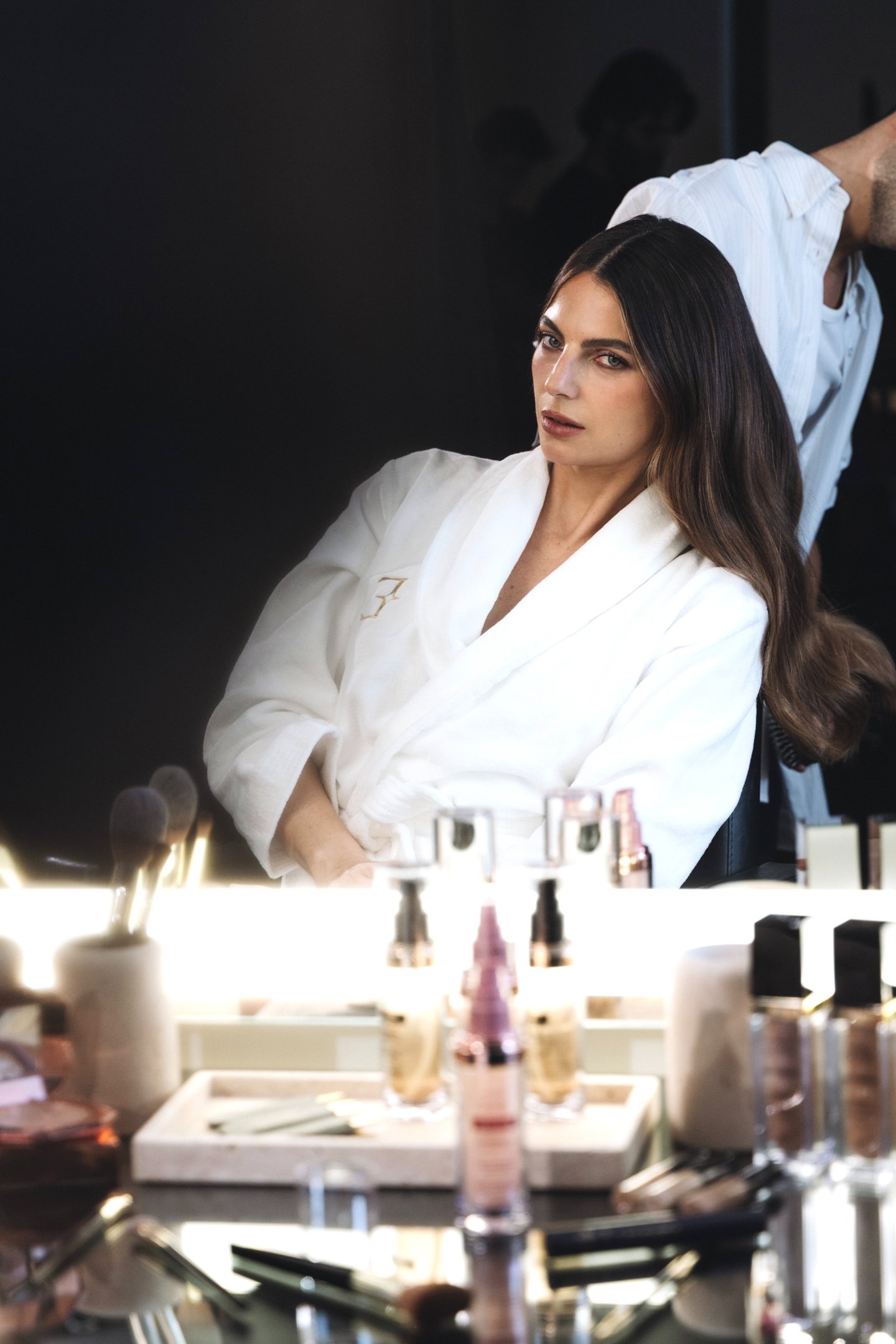 Modelo em roupão branco, sentada em frente a um espelho iluminado com produtos de beleza e maquiagem visíveis. O ambiente reflete um contexto de preparação para um evento de moda, destacando a importância do cuidado com a aparência.