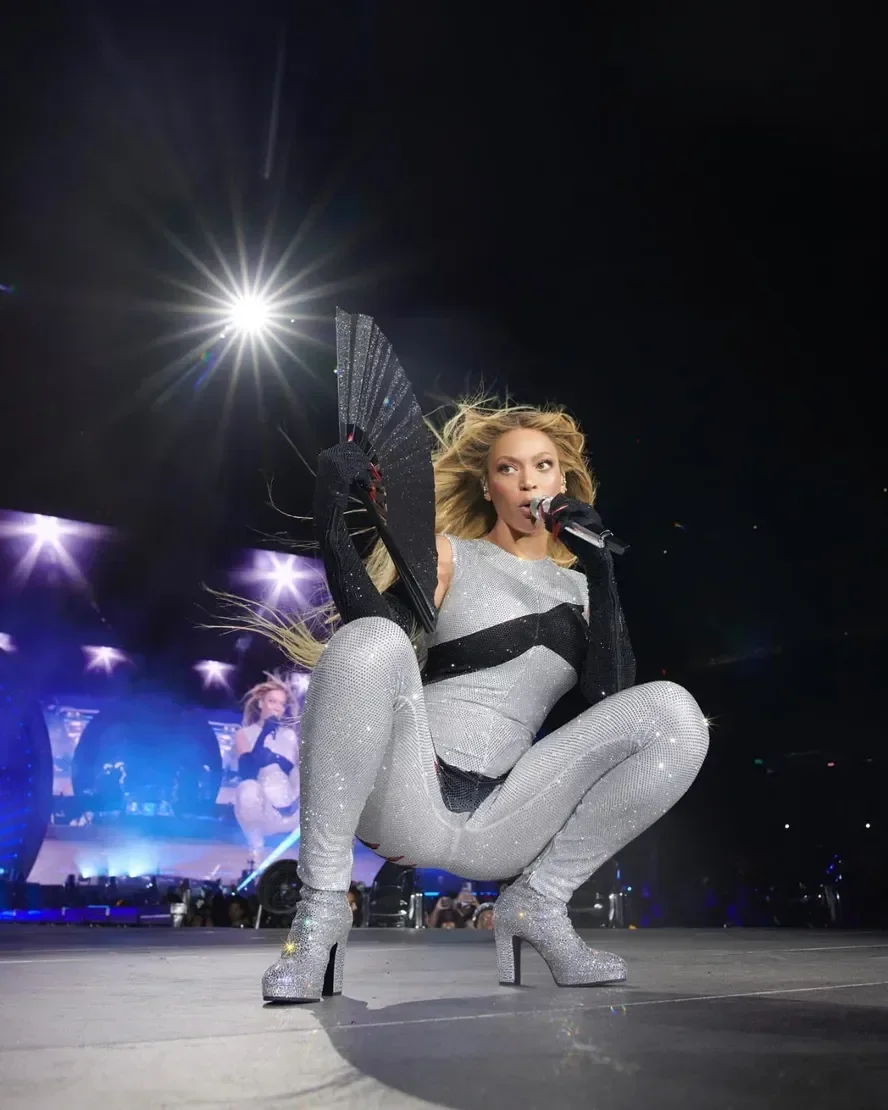 A artista utiliza um leque preto, que complementa seu traje brilhante e ousado. O look, que inclui um macacão prateado e botas de salto, destaca-se em performances de moda e beleza.