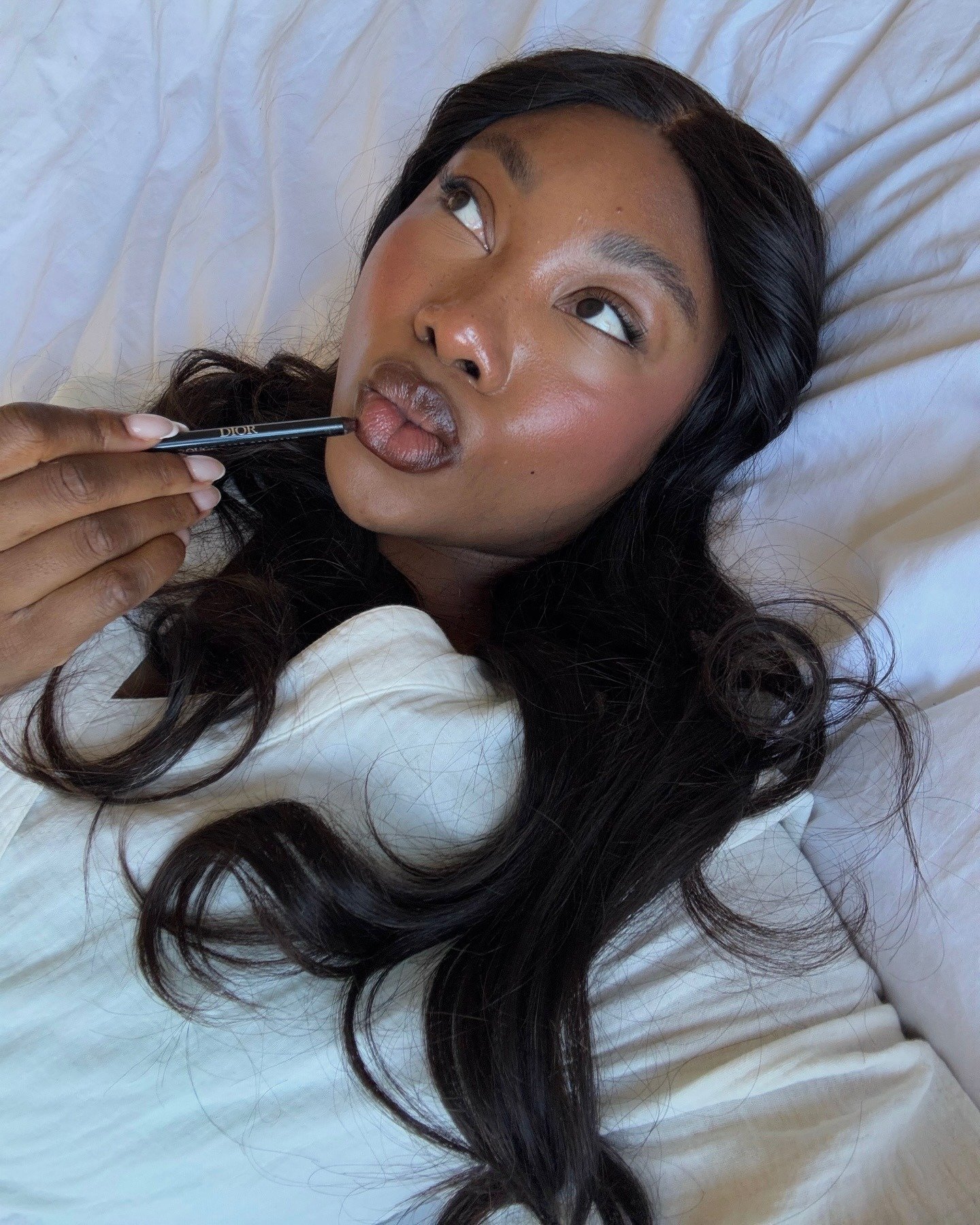 Uma mulher com pele radiante e lábios destacados aplica um lápis labial. Seu cabelo longo e solto emoldura o rosto, enquanto ela observa levemente para o lado. O fundo neutro destaca a beleza e o foco nos produtos de maquiagem.