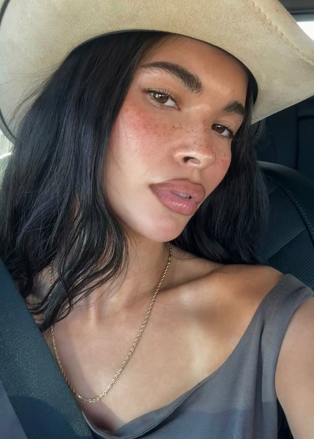 Modelo posa no carro com blush rosa queimado de sol realçando as maçãs, chapéu bege e blusa cinza de tecido leve, clima