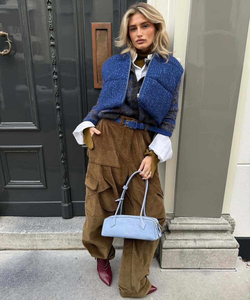 Look moderno com bolsa de ombro estilosa em tom azul claro. Combina peças em camadas: suéter acolchoado, camisa branca e calças de veludo. O visual é complementado por botas vermelhas, trazendo um toque de elegância e conforto.