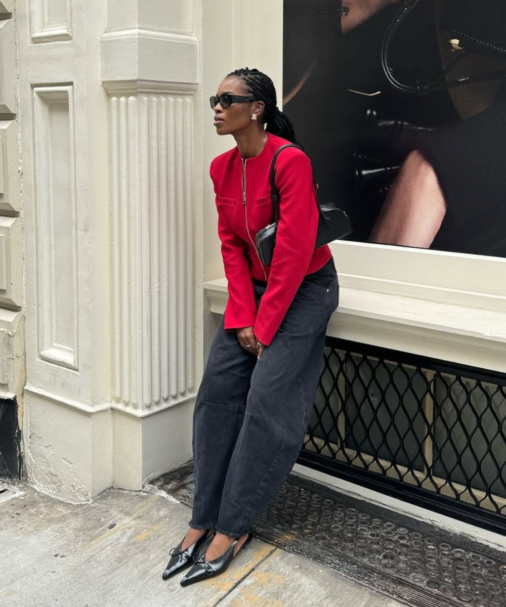 Mulher com bolsa de ombro estilosa, usando blazer vermelho e calças largas pretas, destaca-se em cenário urbano moderno. Óculos escuros e sapatos elegantes completam o look.