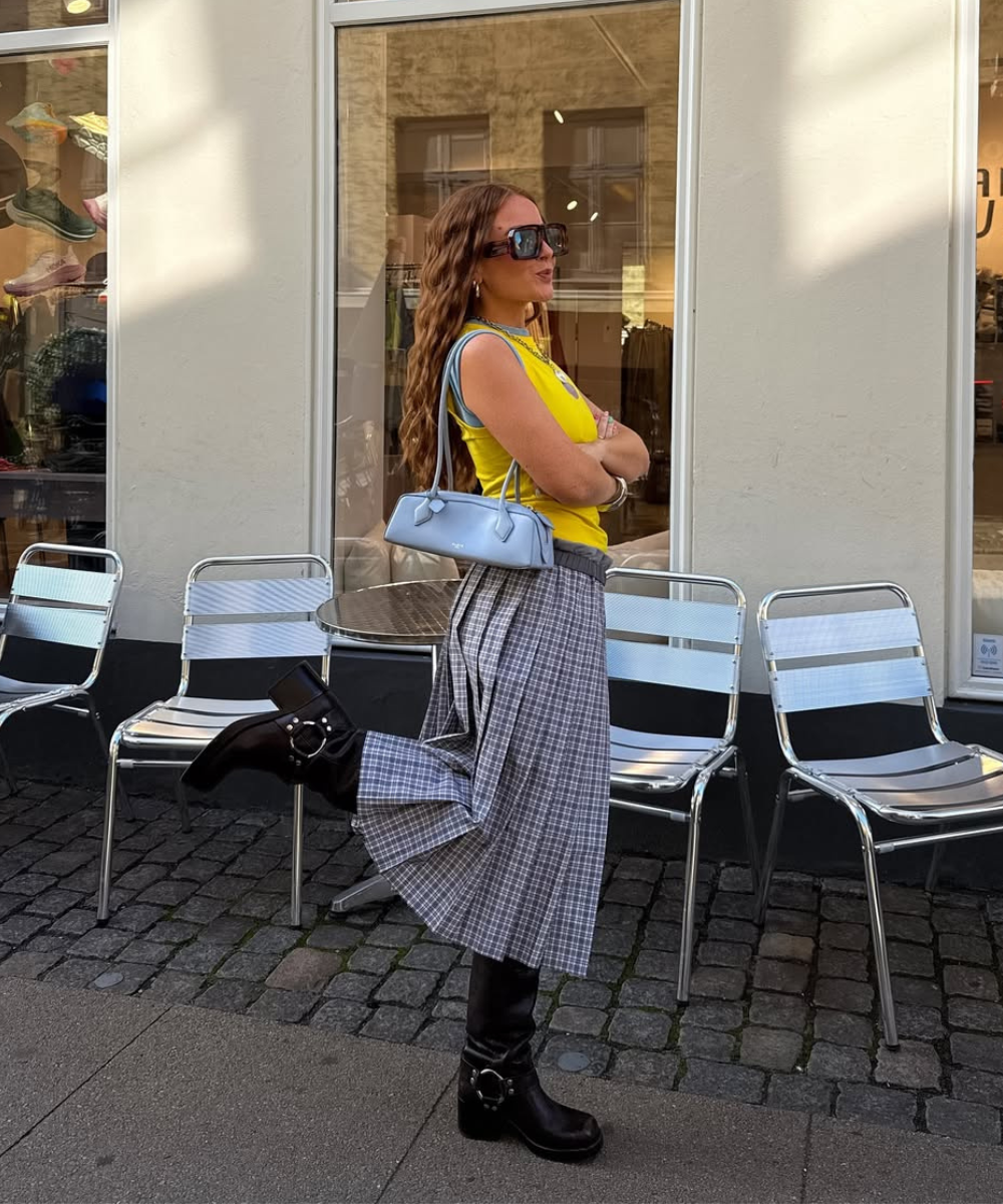 Mulher com bolsa de ombro estilosa em tom claro, usando blusa amarela e saia xadrez. Botas pretas robustas completam o look. Ideal para quem busca inspiração em moda contemporânea.