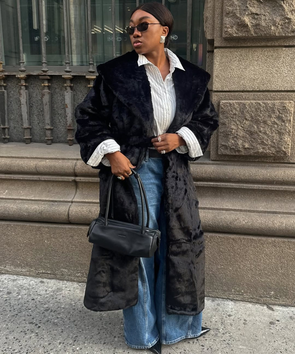 A imagem apresenta uma mulher elegante com uma bolsa de ombro estilosa em preto, combinando perfeitamente com seu casaco de pelúcia. A blusa listrada e o jeans amplo completam o look, destacando a sofisticação e o conforto.