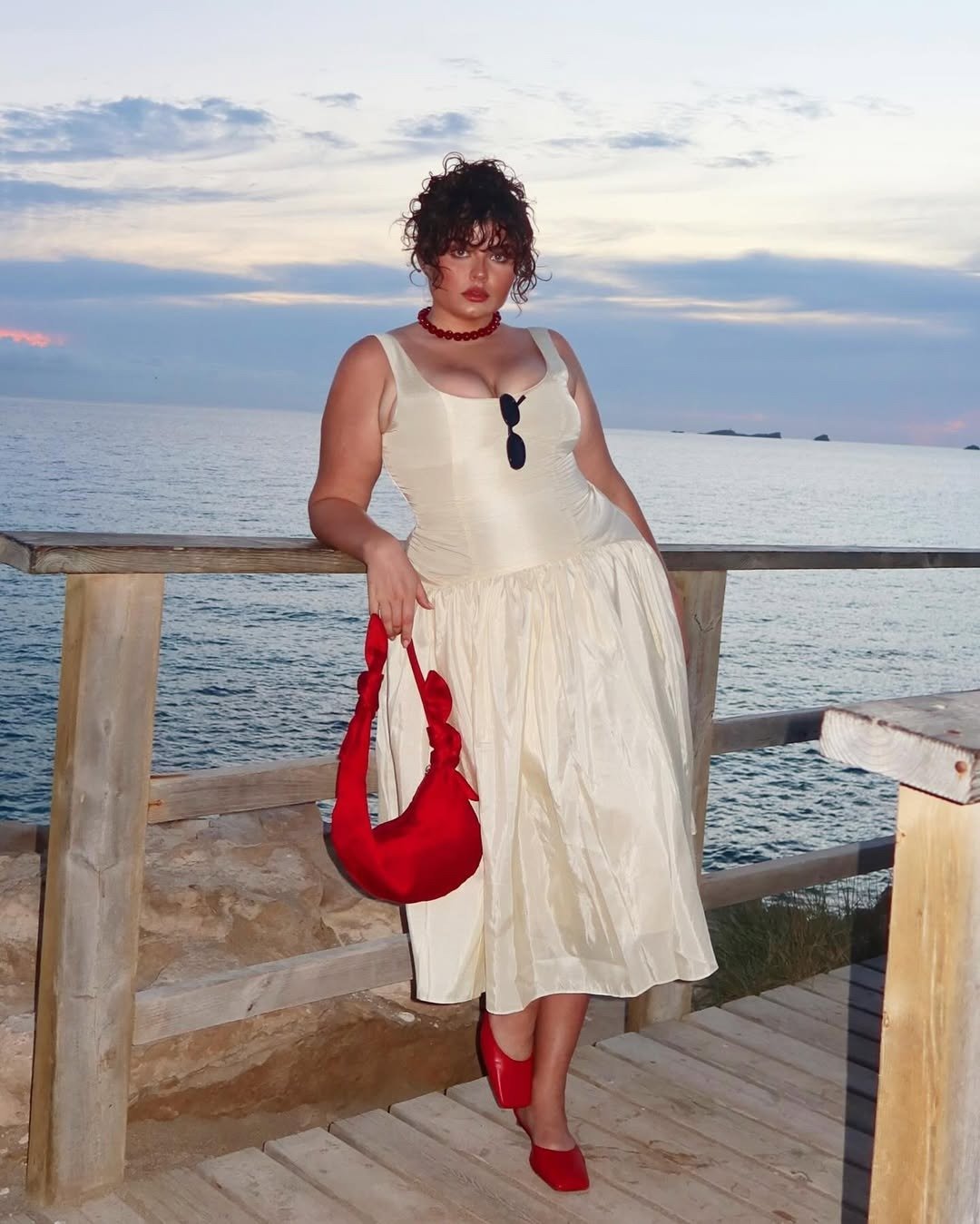 Modelo posa à beira-mar com vestido midi off-white de tecido leve e estruturado, combinando bolsa vermelha de alça torcida.