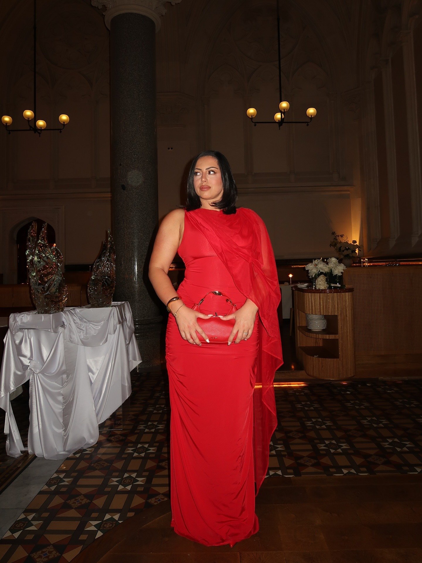 Mulher posa com vestido longo vermelho justo e tecido fluido, segurando bolsa vermelha de couro com alça de argolas.