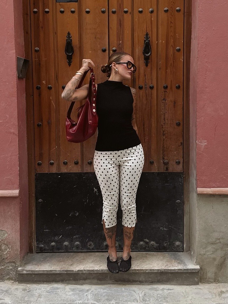 Modelo posa segurando bolsa vermelha de couro com detalhes em botão, combinada a regata preta e calça branca de bolinhas