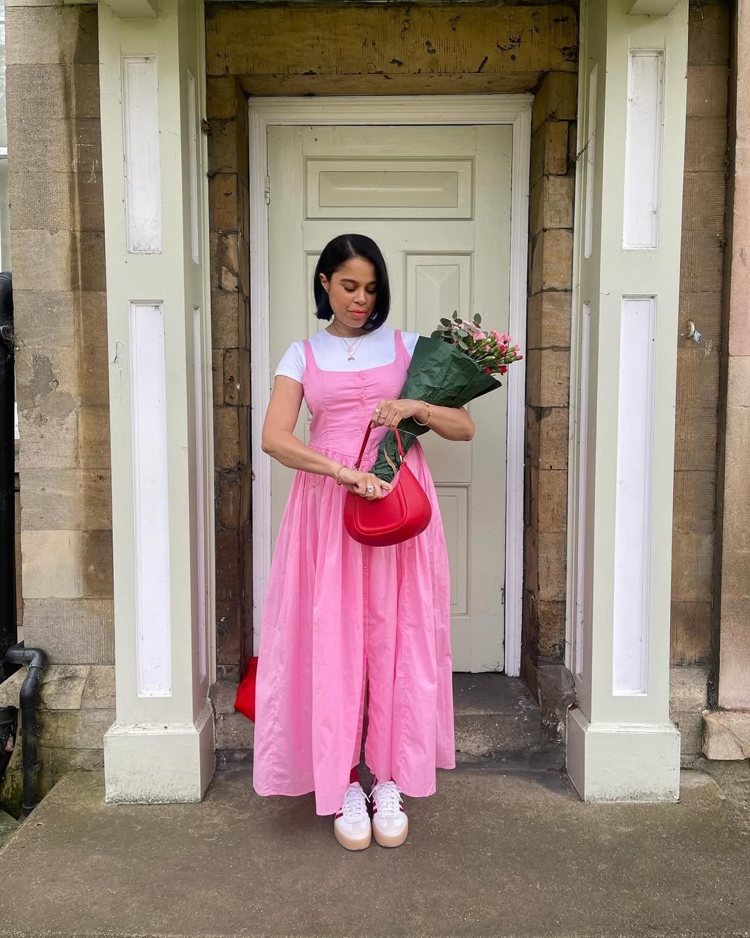 Mulher segura buquê e ajusta bolsa vermelha de couro com vestido rosa fluido e camiseta branca, em clima delicado e casual.
