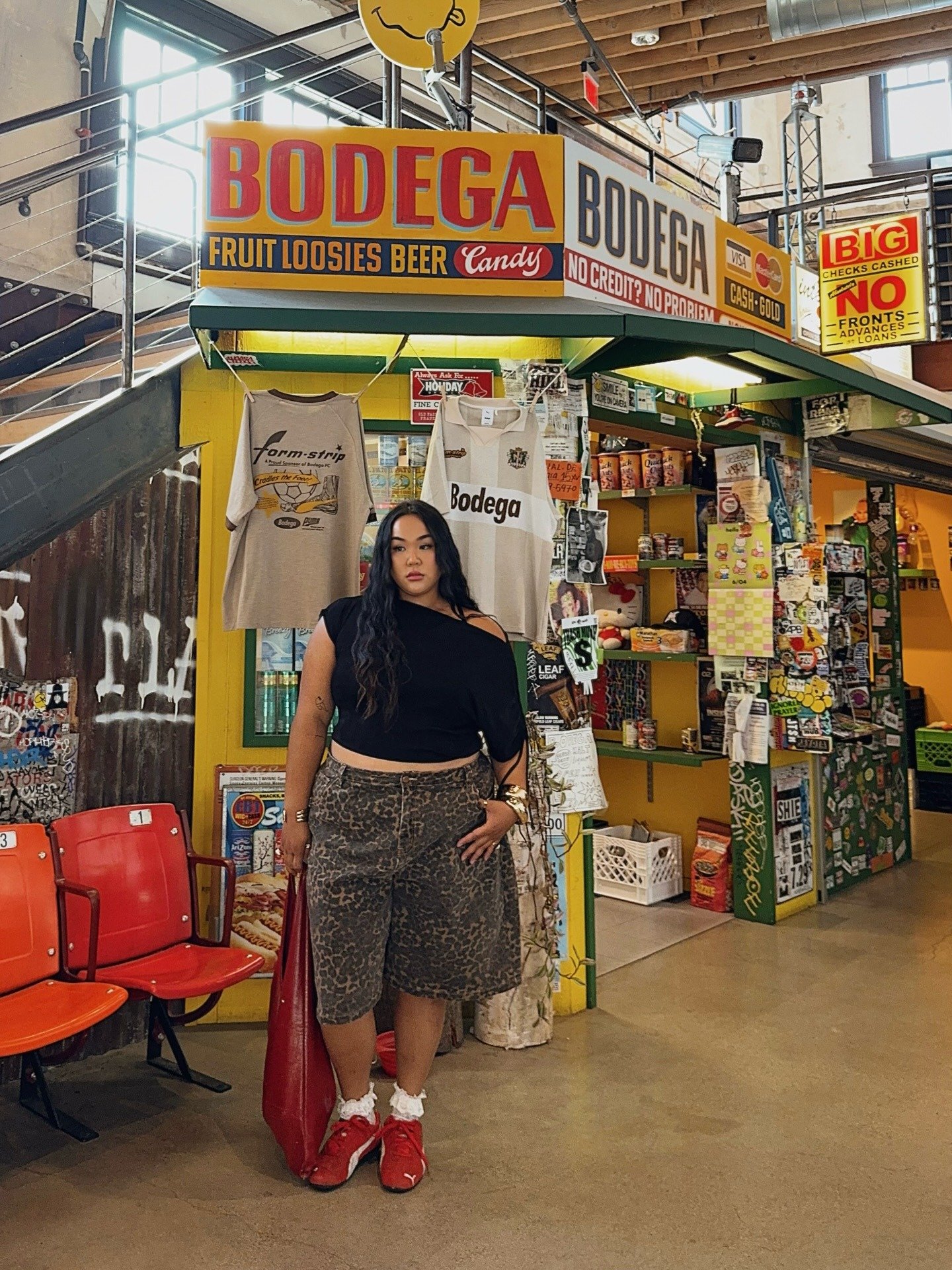 Modelo posa com blusa preta de ombro só, bermuda animal print e tênis vermelho, segurando bolsa vermelha de couro grande.