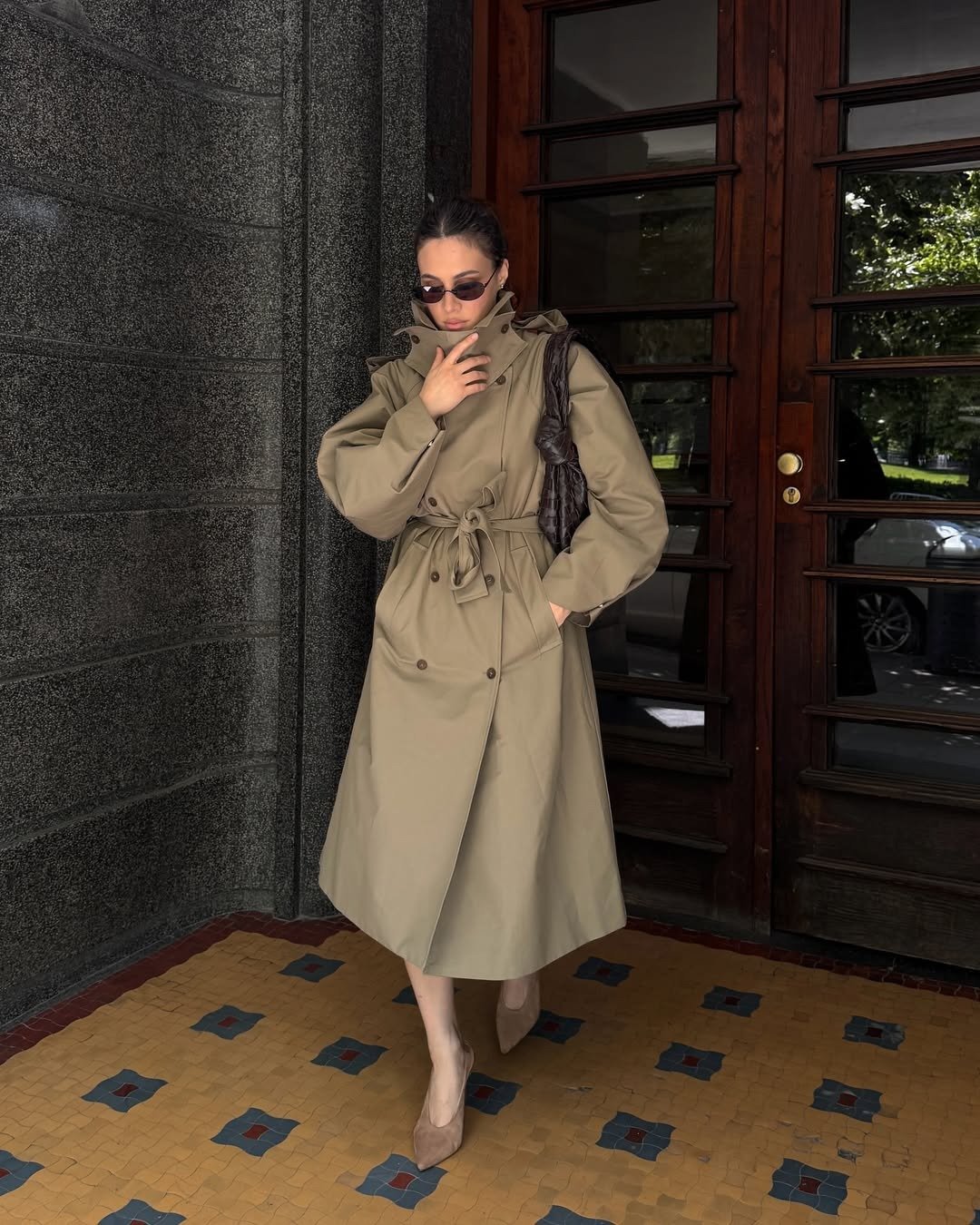 Modelo posa com trench coat bege de tecido estruturado, gola funil alta e amarração na cintura, combinando scarpin nude e