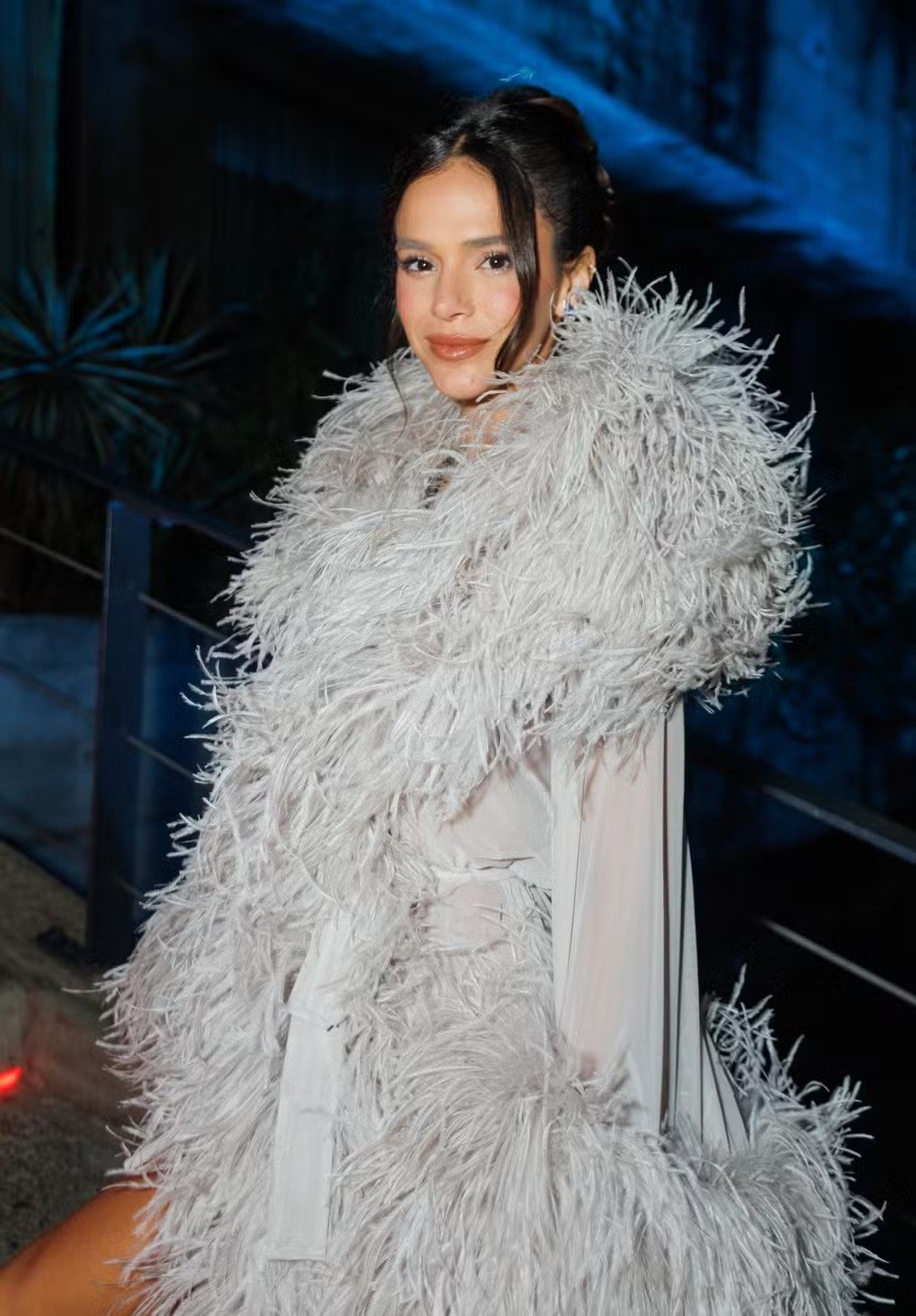 Bruna Marquezine exibe um visual impactante com um casaco de penas cinza claro, destacando-se em um ambiente iluminado.