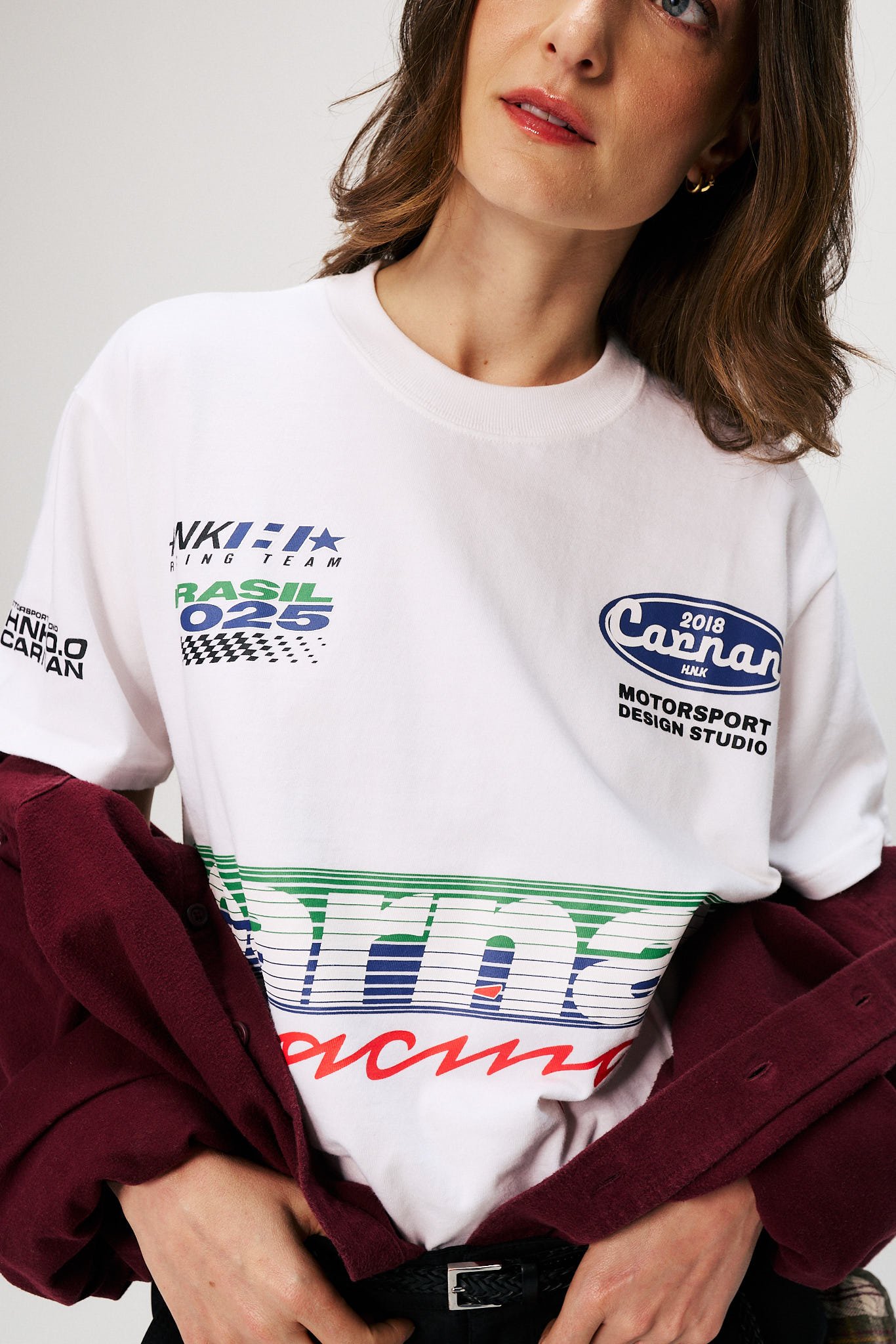 Uma mulher veste uma camiseta branca com estampas coloridas, incluindo referências a corridas e design de moda. Ela segura uma camisa de flanela marrom, que complementa o look.