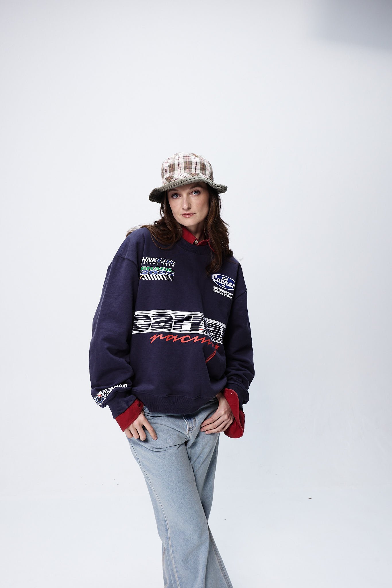 A modelo exibe um look moderno e despojado, combinando um moletom oversized azul escuro com detalhes coloridos e uma camisa vermelha por baixo. O uso de um chapéu bucket xadrez complementa o visual.