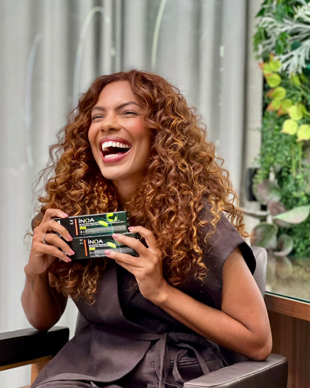 Modelo posa com coloração de cabelos crespos e cacheados loiro acobreado vibrante, roupão cinza.