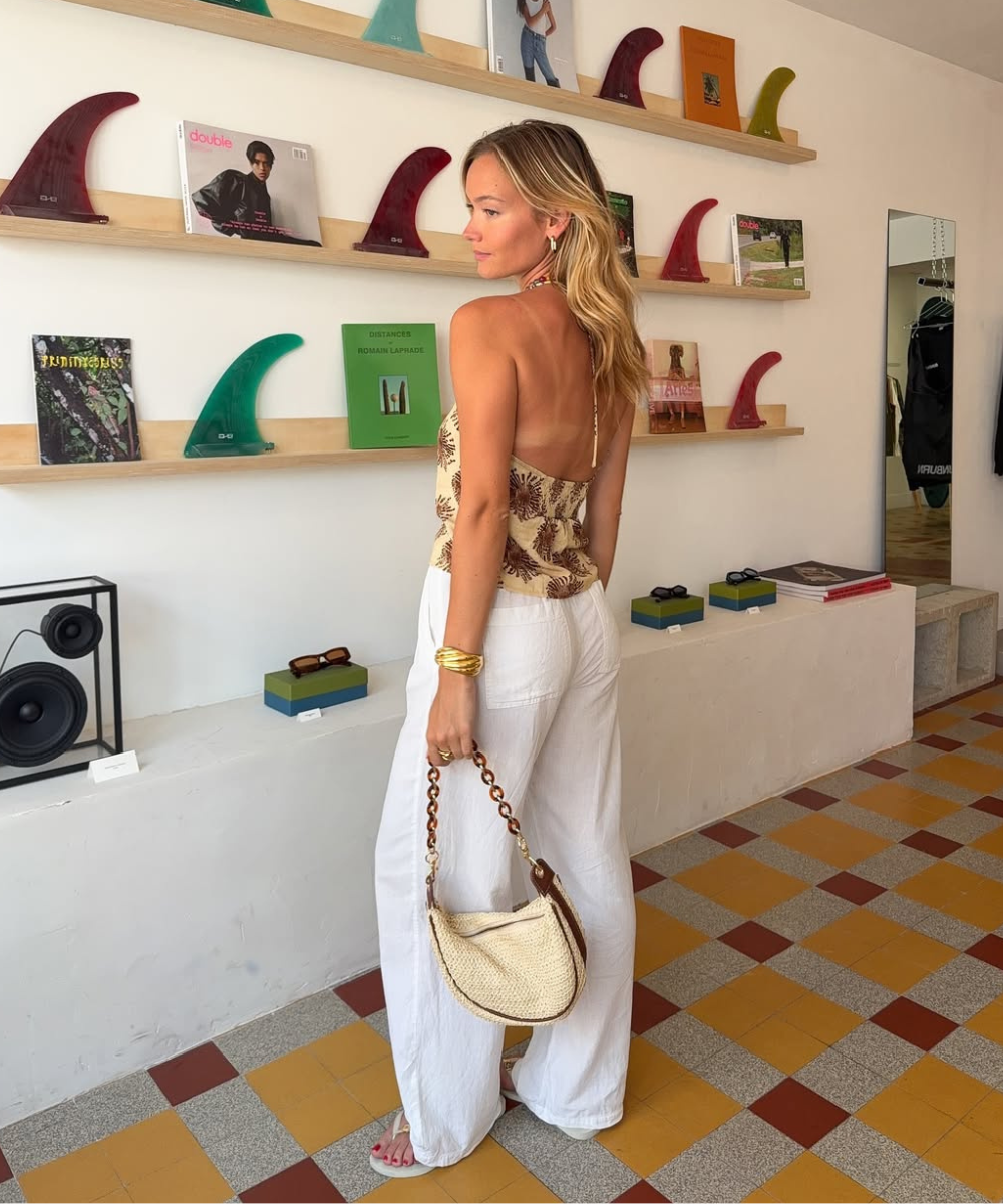 Modelo posa de costas com calça de linho branca ampla, blusa bege estampada e bolsa de palha, em ambiente moderno e