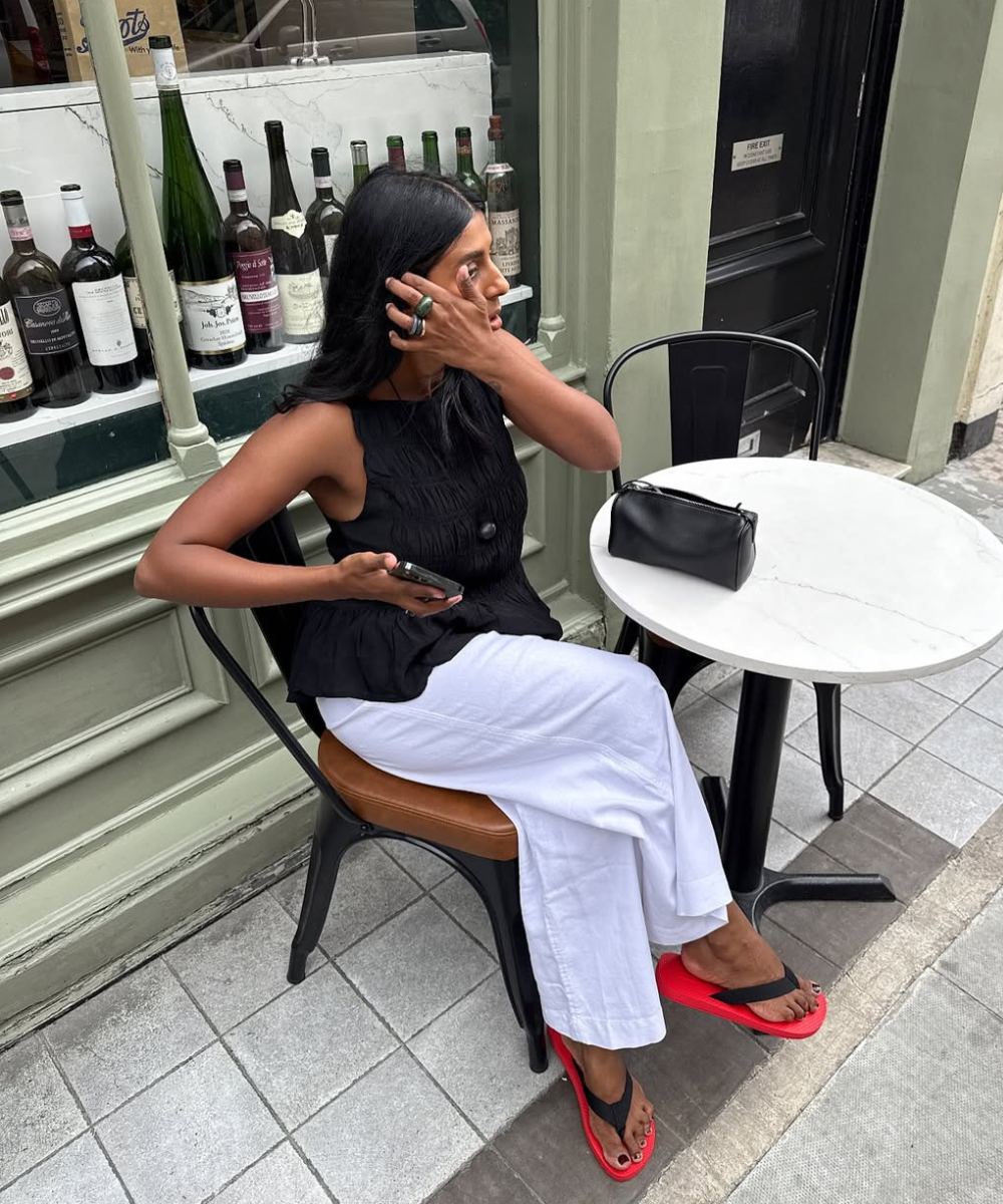 Mulher sentada em cadeira preta com calça de linho branca reta, blusa preta texturizada e chinelos vermelhos, em ambiente