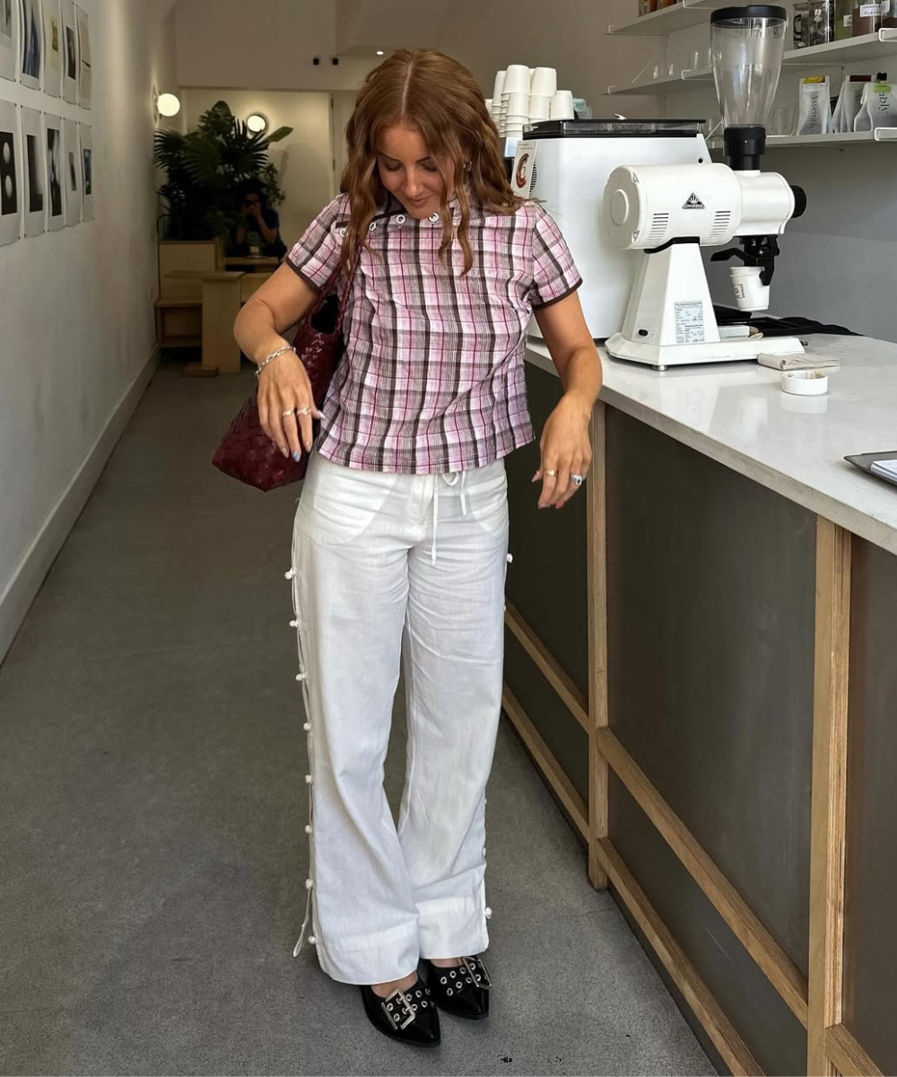 Modelo posa olhando para baixo com calça de linho branca ampla com botões laterais, blusa xadrez rosa e sapatos pretos em