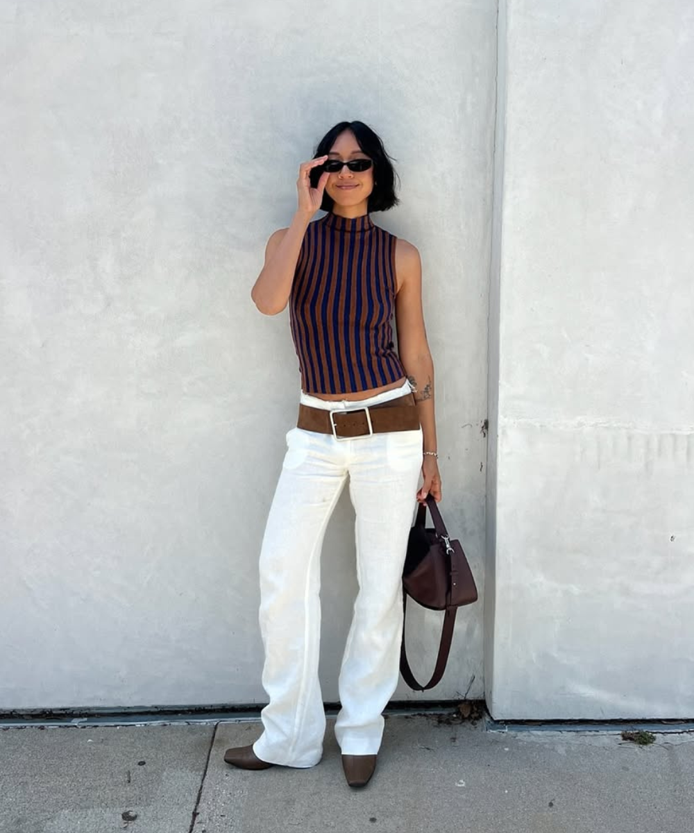Modelo posa com calça de linho branca reta, blusa regata listrada marrom e azul, cinto largo marrom e bolsa combinando, em