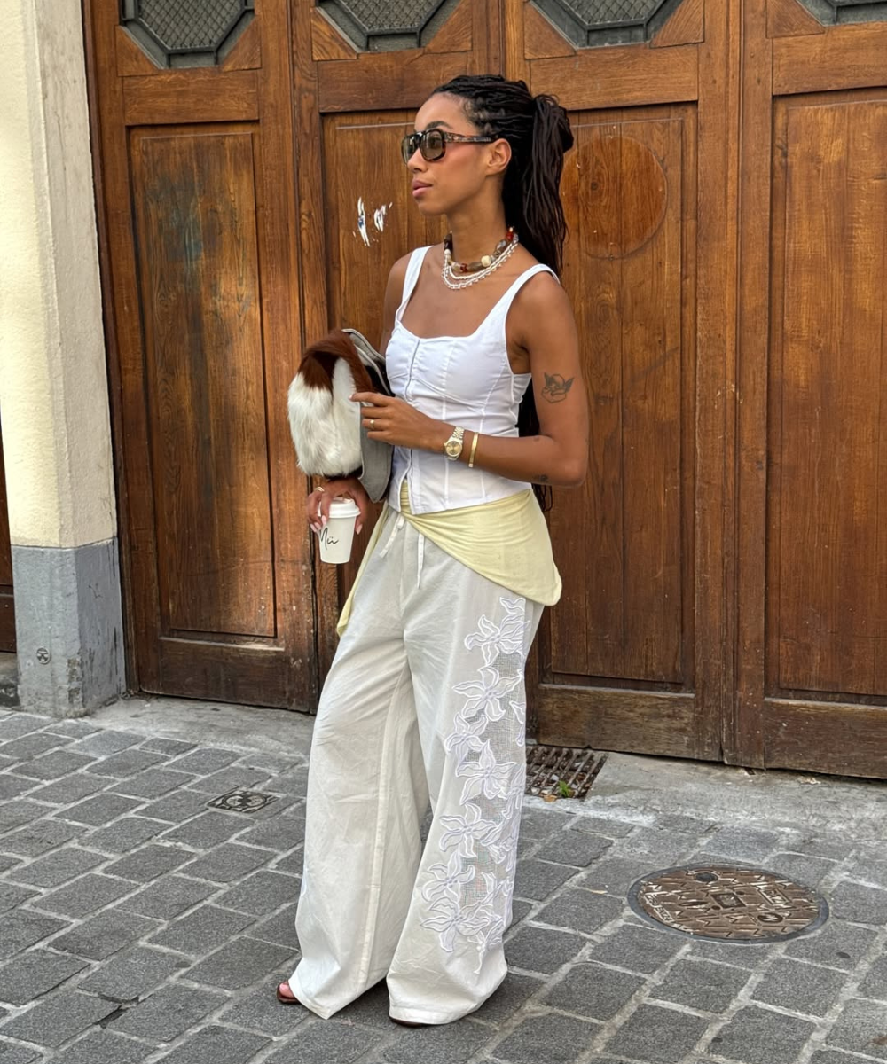 Modelo posa de lado com calça de linho branca ampla e bordada, regata branca justa e acessórios neutros em ambiente urbano.