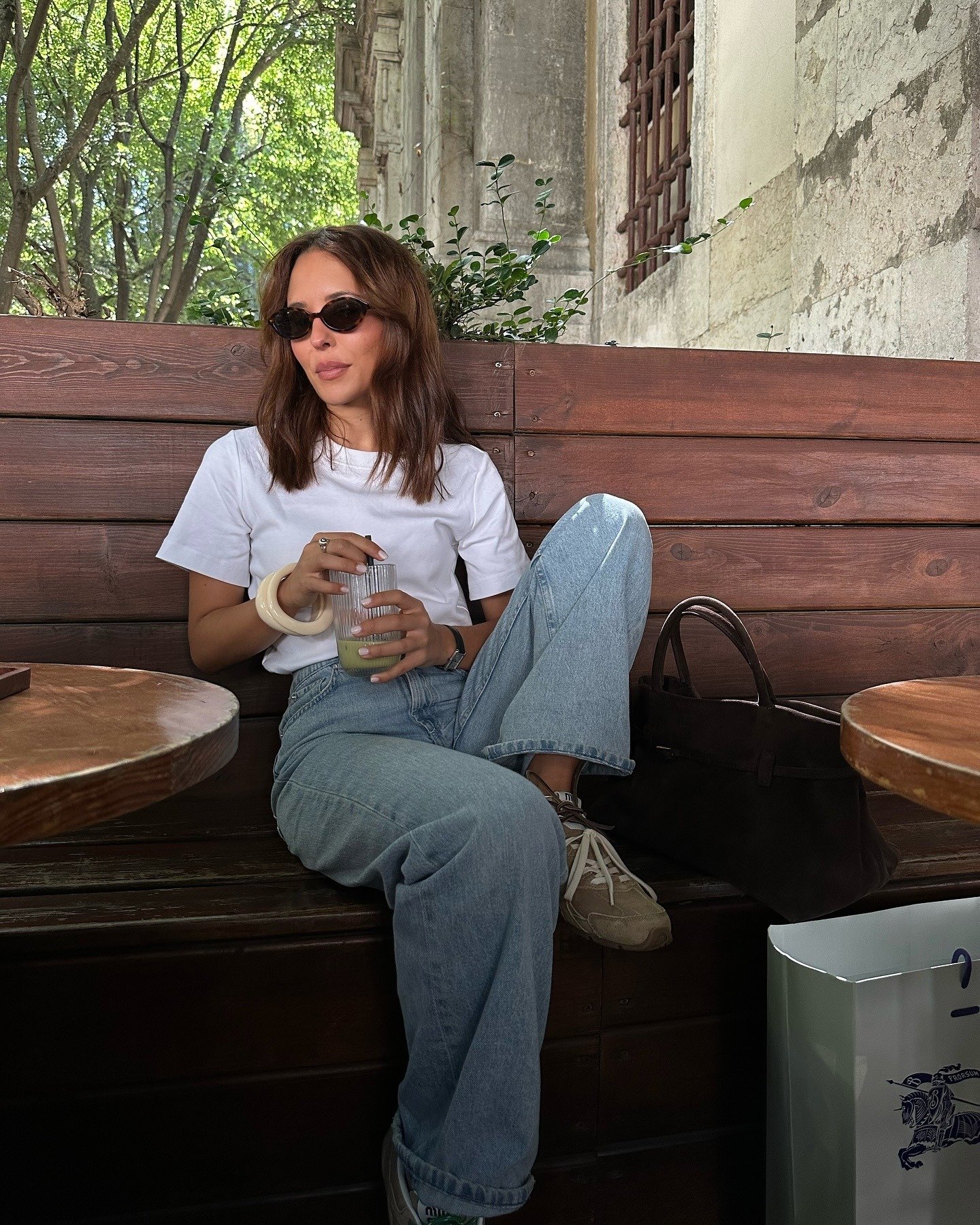 Mulher sentada com calça jeans e tênis bege, camiseta branca e acessórios neutros em ambiente rústico e