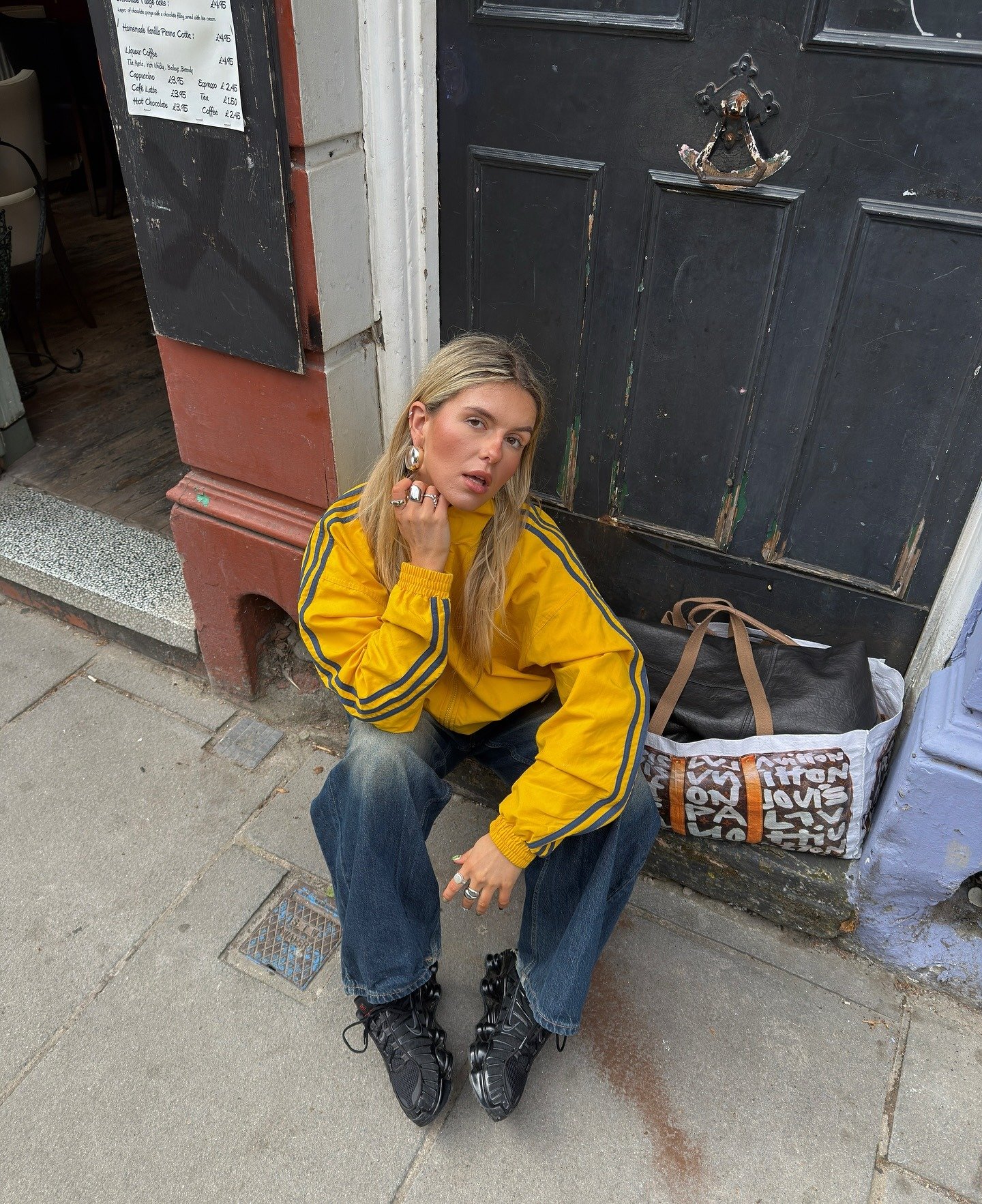 Modelo senta no chão com calça jeans ampla e tênis preto robusto, combinando jaqueta amarela vibrante com listras cinza, em