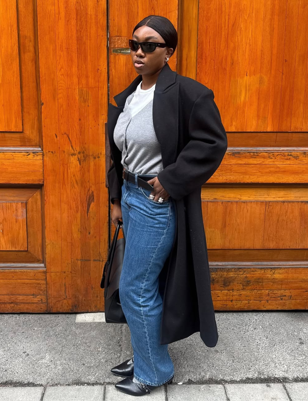 A imagem apresenta uma mulher vestindo calças jeans de cintura alta, uma tendência forte para 2026. As calças têm um corte reto, combinando com uma camiseta cinza e um casaco preto oversized, que traz sofisticação ao look.