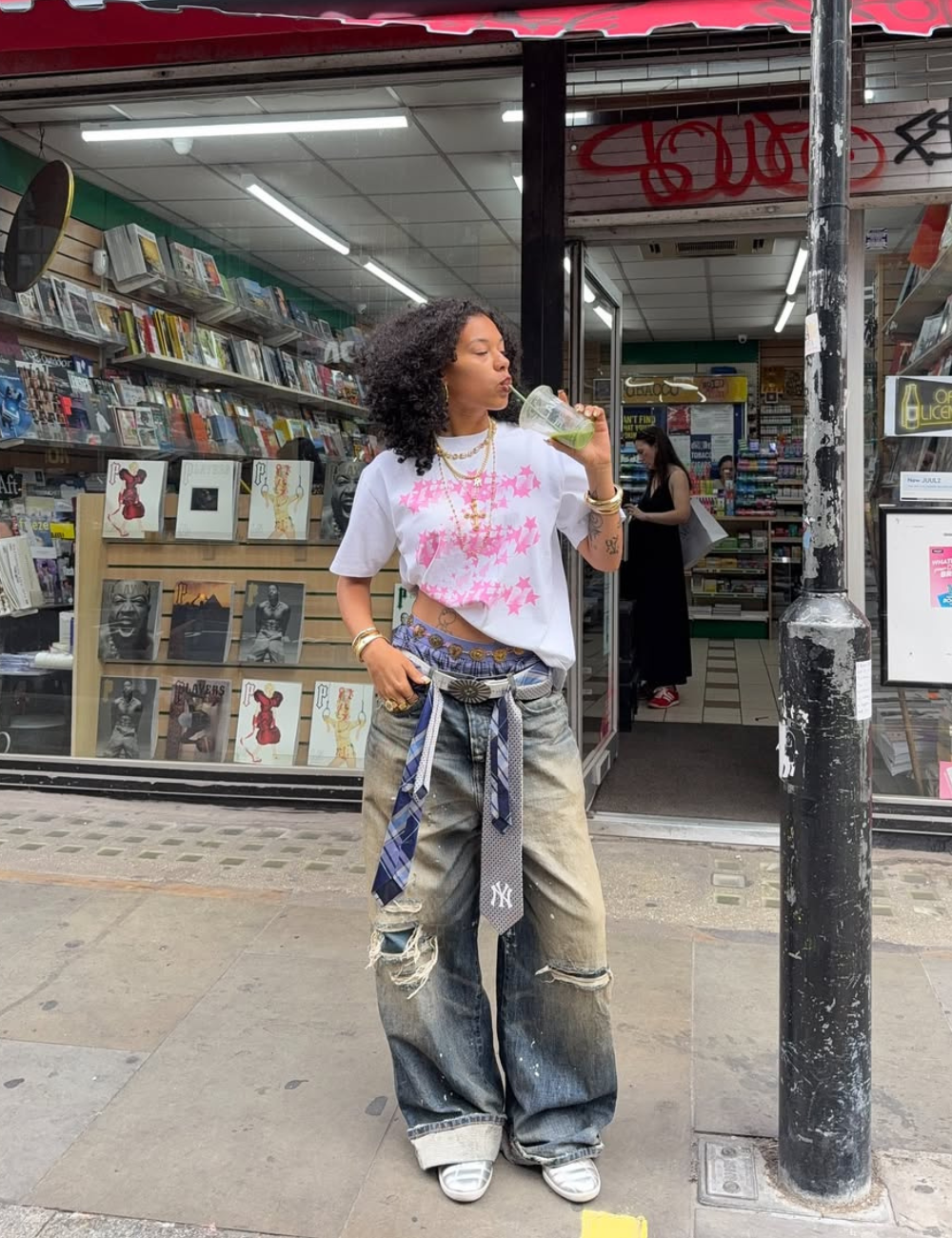 A imagem apresenta uma mulher estilosa usando calças jeans oversized, tendência para 2026. As calças têm detalhes desgastados e são acompanhadas por uma camiseta branca com estampa rosa.