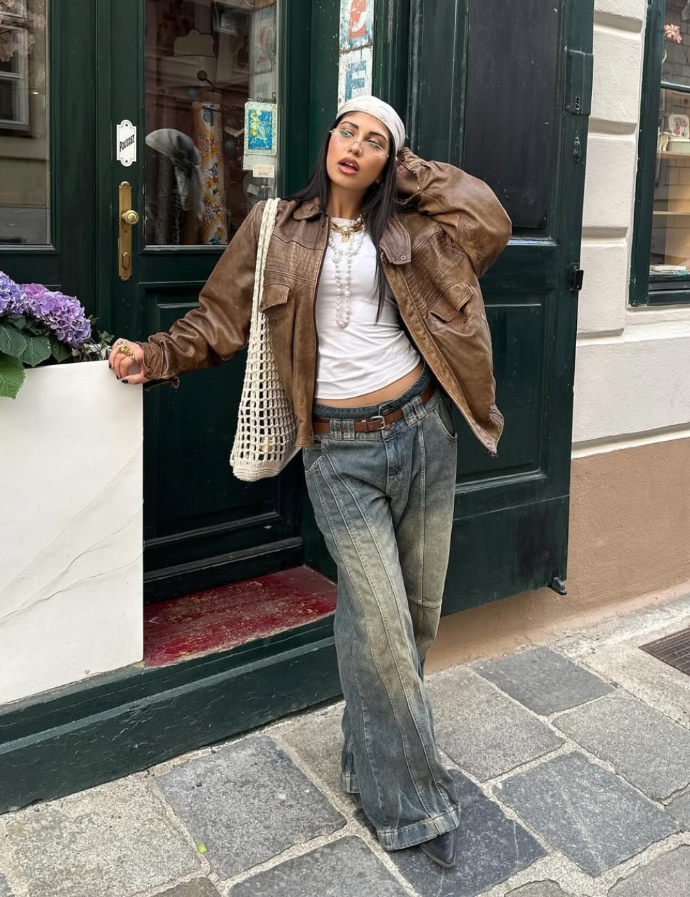 A imagem apresenta uma jovem estilosa, usando calças jeans largas com um efeito desbotado, tendência forte para 2026. Ela combina a peça com uma blusa branca básica e uma jaqueta de couro marrom, que traz um toque vintage.