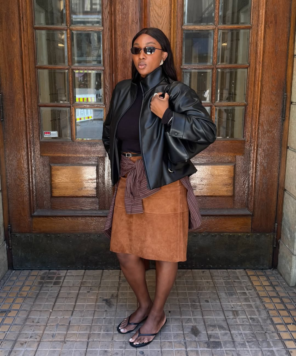 Look moderno com camisa amarrada na cintura, combinando jaqueta de couro, blusa preta e saia de camurça. Óculos escuros adicionam estilo.