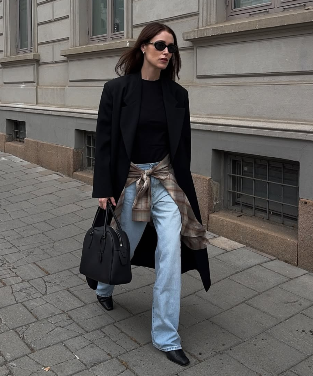 Look moderno com camisa amarrada na cintura, combinando blazer oversized e jeans claros. Óculos escuros e bolsa preta completam o visual. Essa combinação é ideal para um estilo casual e elegante, trazendo versatilidade e conforto.