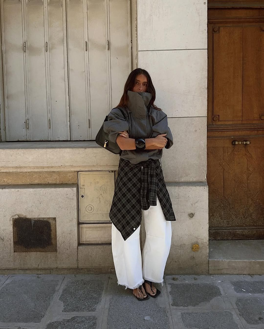 Modelo posa com postura confiante usando blusa cinza de gola funil estruturada, calça branca ampla e camisa xadrez preta