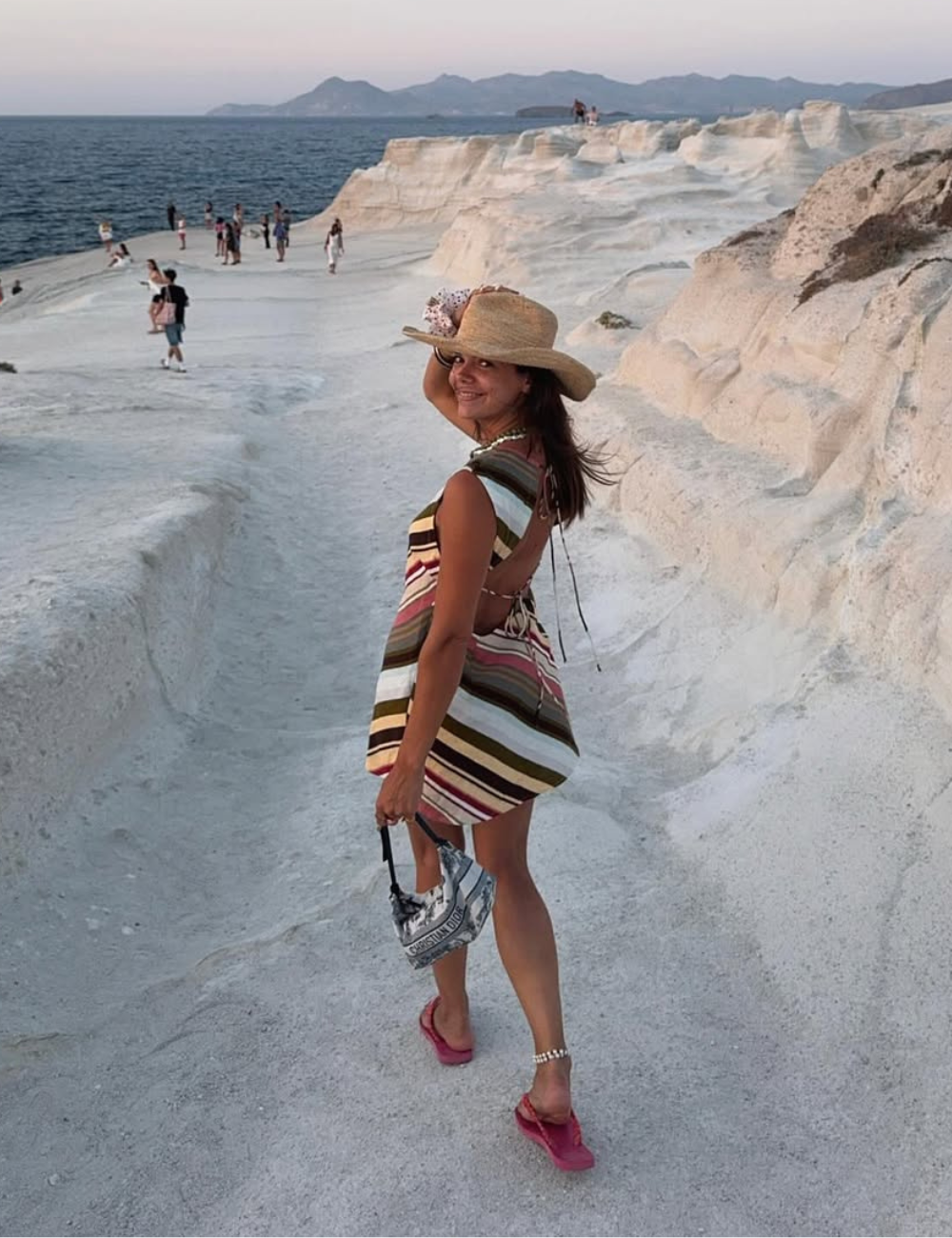 Um chapéu de palha elegante complementa o visual de uma mulher sorridente em um cenário à beira-mar. Ela usa um vestido listrado em tons terrosos, que destaca sua silhueta.