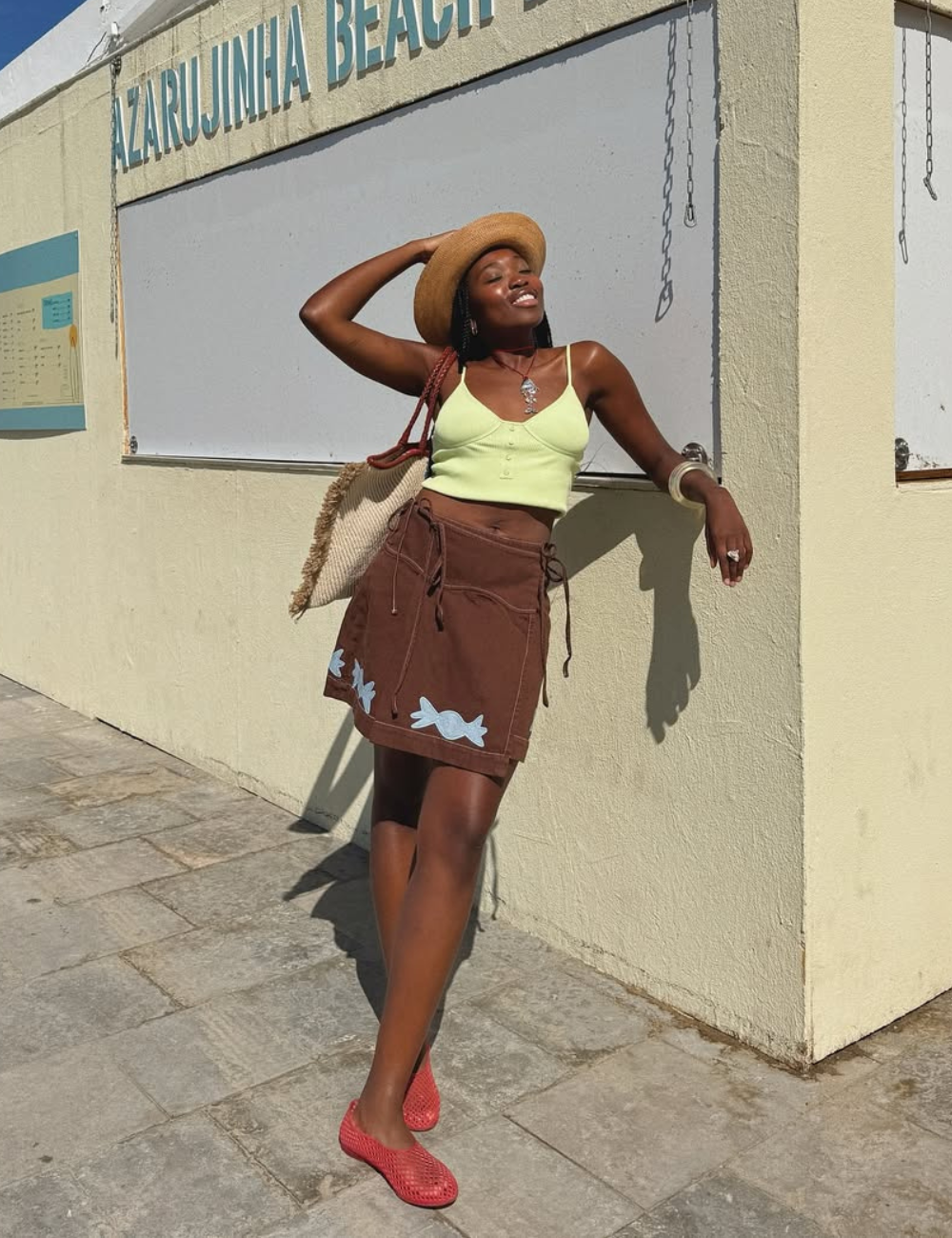 O chapéu de palha complementa perfeitamente o look leve e descontraído, ideal para dias ensolarados. A combinação de um top verde claro e uma saia marrom com detalhes em azul cria um visual harmonioso.