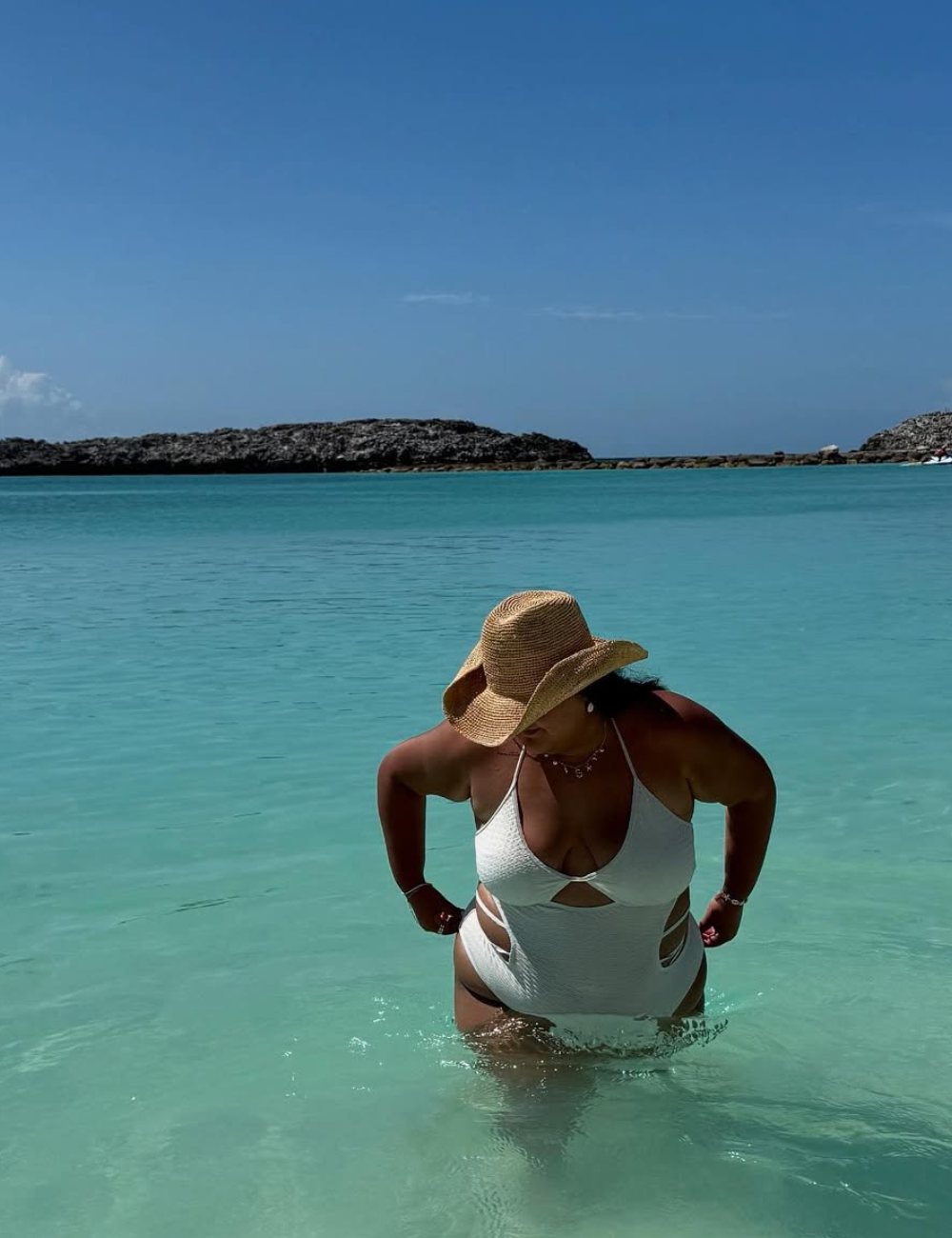 Um chapéu de palha elegante complementa o visual de banho em uma praia paradisíaca. A modelo, vestindo um maiô branco com recortes, está em águas cristalinas, refletindo o estilo de verão.