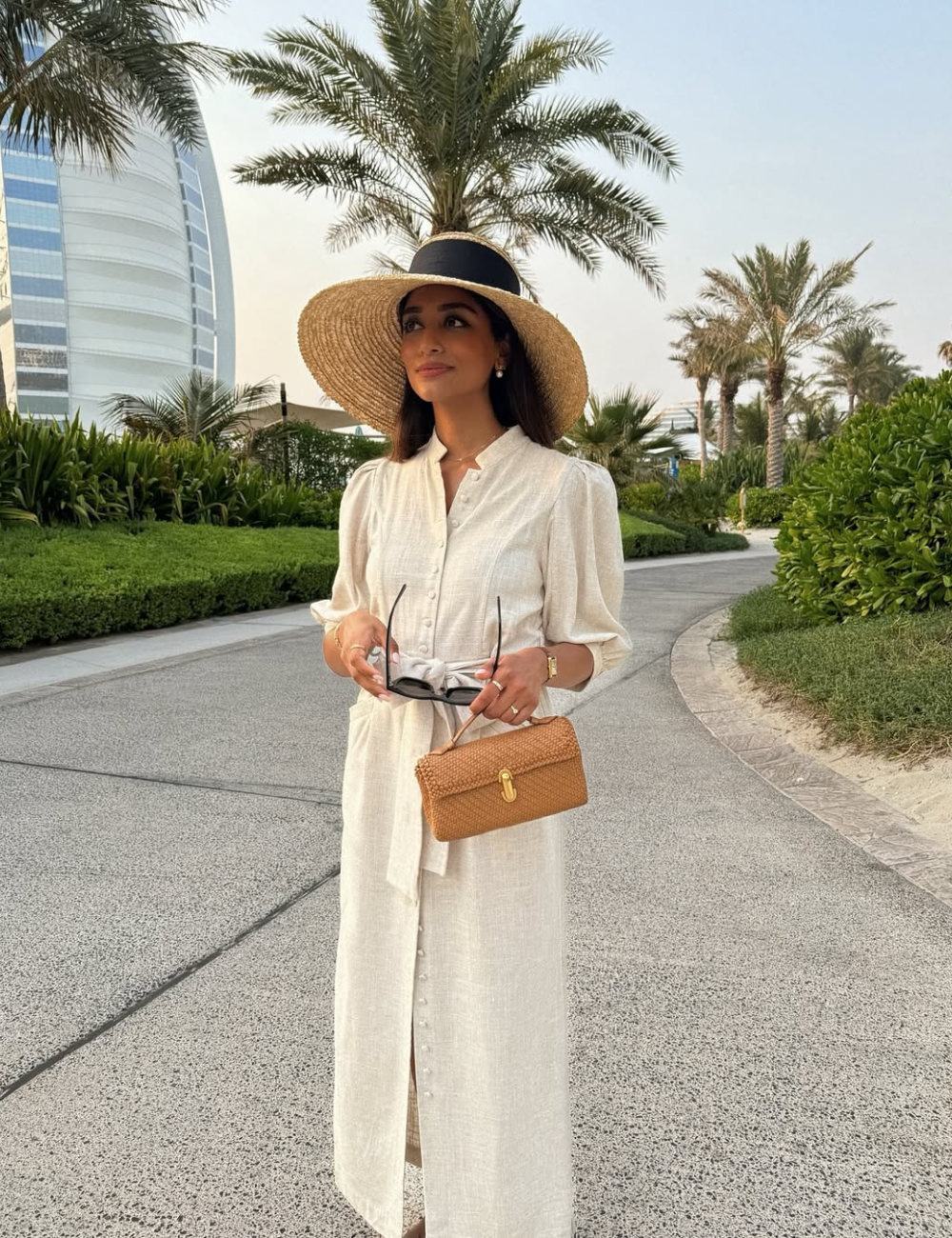 Com um elegante chapéu de palha, a mulher exibe um visual sofisticado. O chapéu, de aba larga e detalhe em preto, complementa um vestido longo de linho claro, com botões frontais e um cinto na cintura, ideal para dias ensolarados.