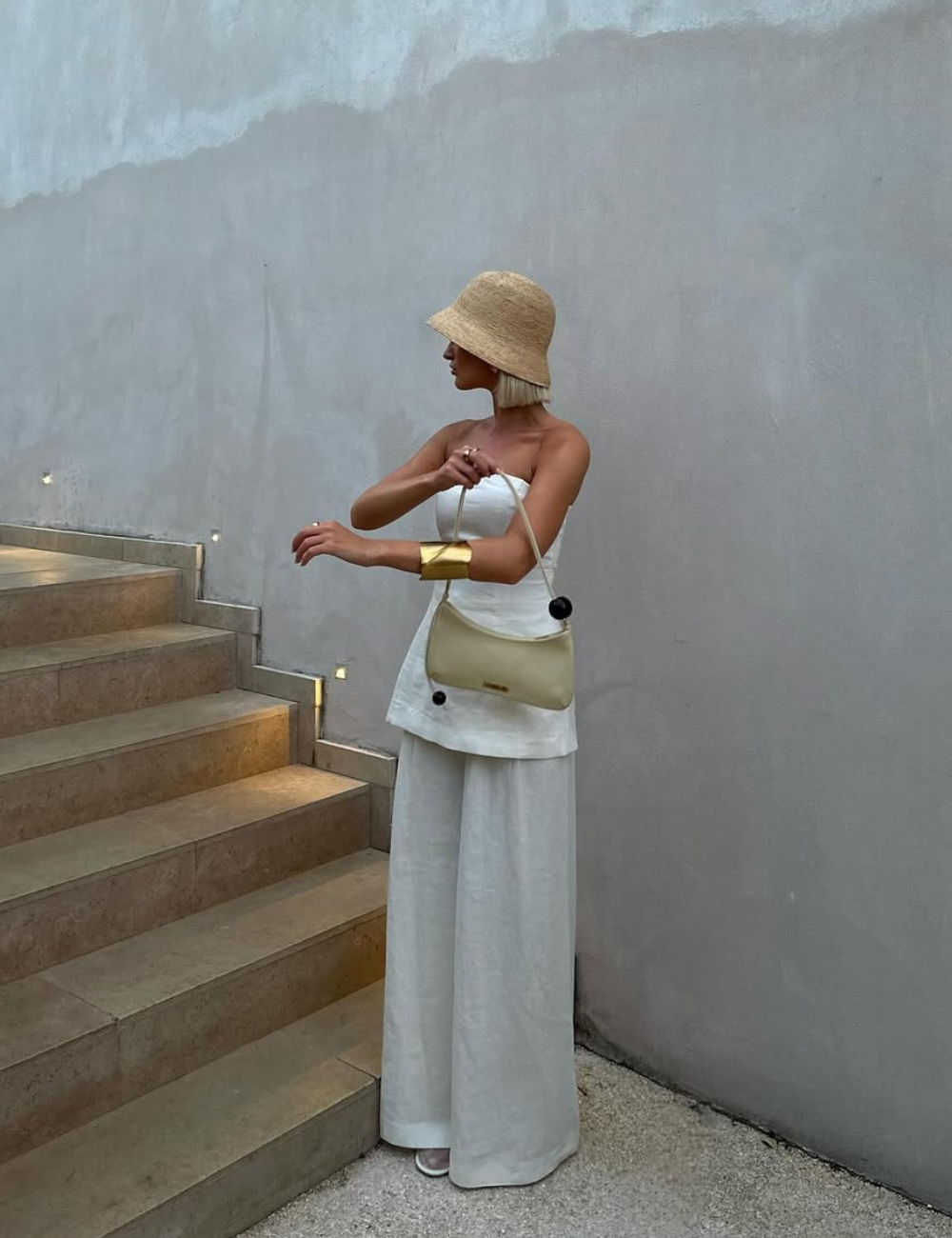 O chapéu de palha, com sua forma estilizada, complementa o look leve e elegante. A modelo usa uma blusa branca e calças largas em tom claro, criando um visual fresco ideal para dias quentes. A bolsa amarela adiciona um toque moderno.