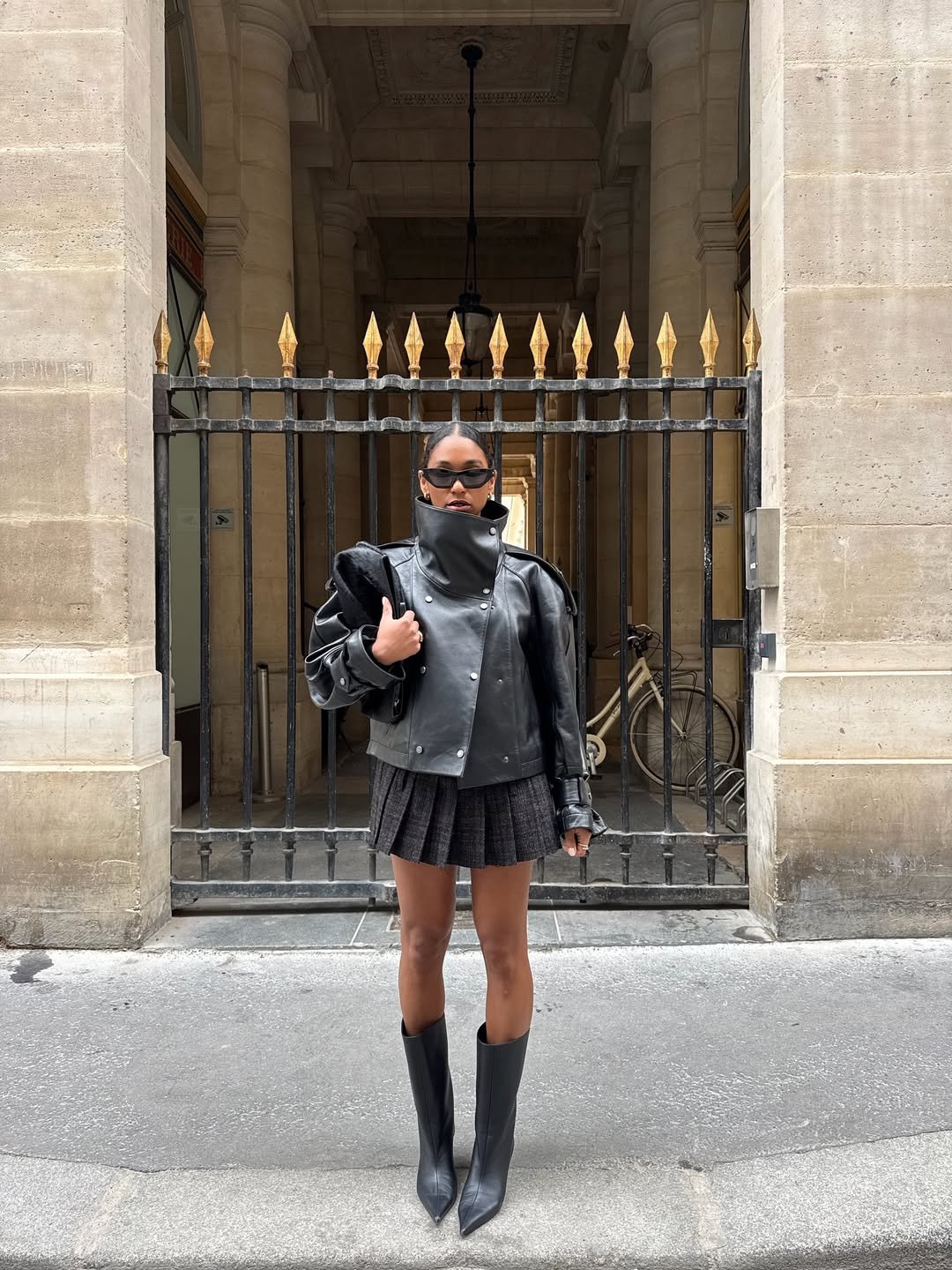 Modelo posa em jaqueta de couro preta com gola funil alta, saia plissada cinza e botas longas, em clima urbano sofisticado.