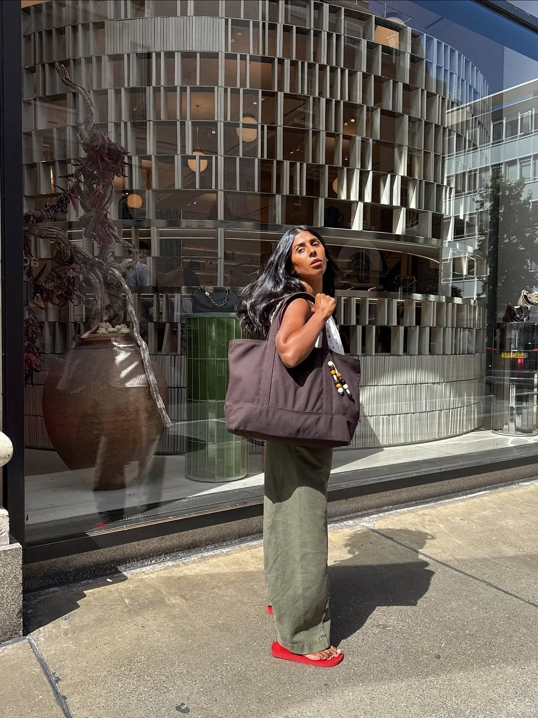 Mulher posa ao sol com saia longa verde, blusa branca e chinelo vermelho vibrante, carregando bolsa marrom oversized.