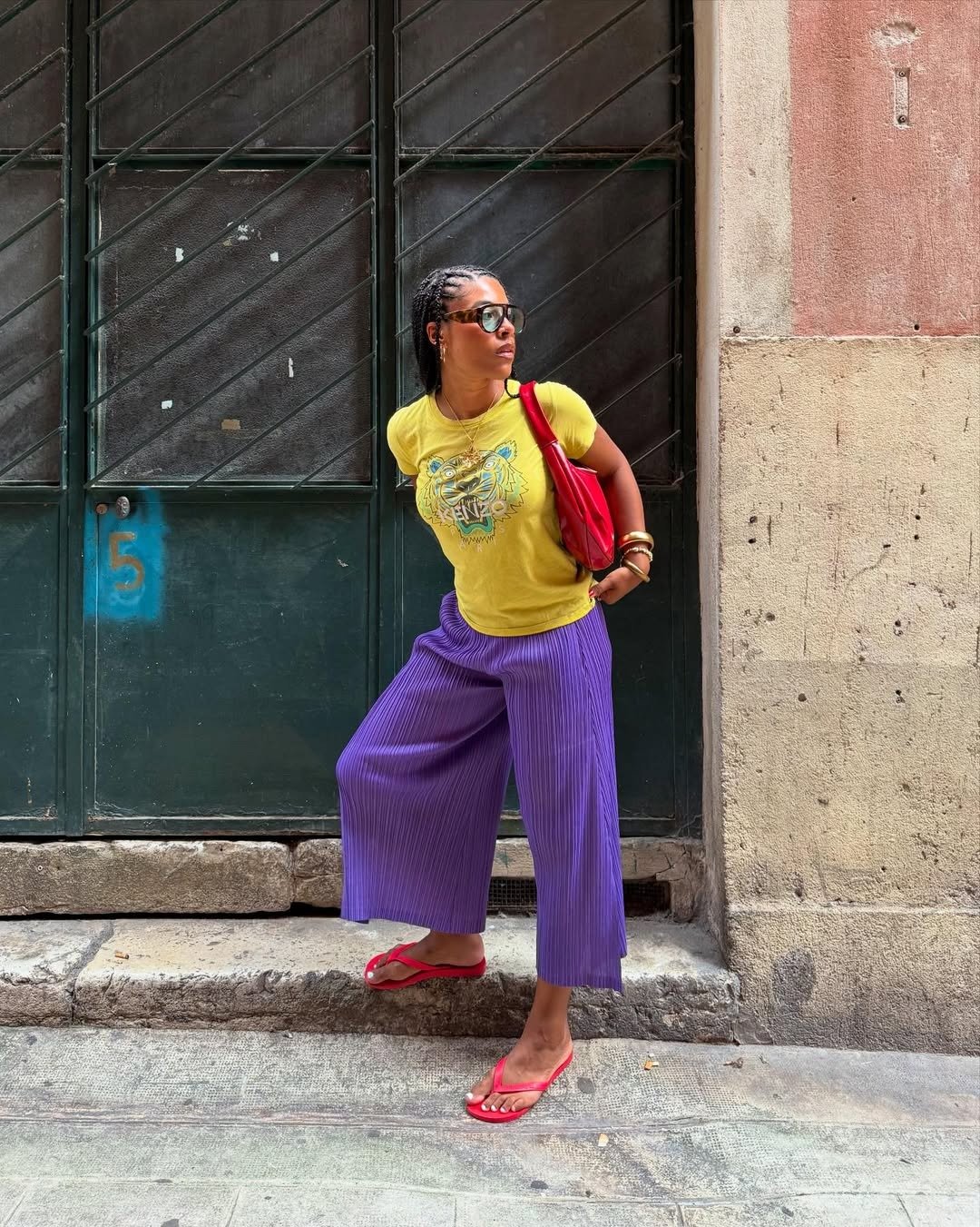 Modelo posa com postura confiante usando chinelo vermelho de tiras finas, calça roxa ampla plissada e camiseta amarela.