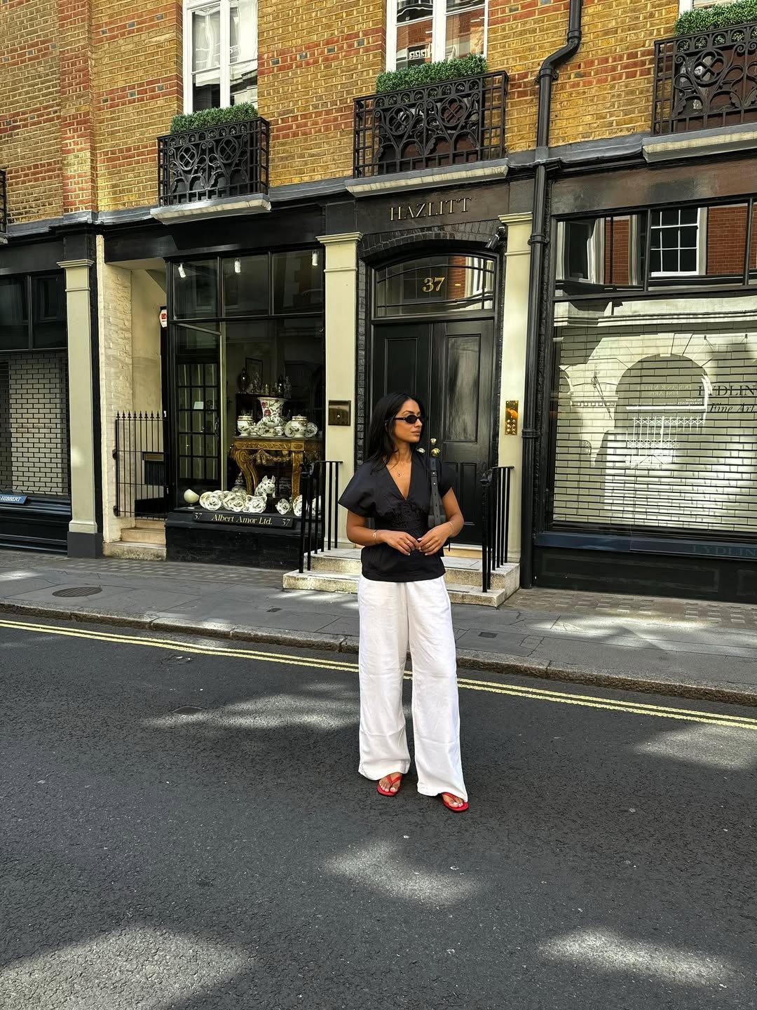 Modelo posa na rua com blusa preta de mangas bufantes, calça branca ampla e chinelo vermelho de tiras finas, em look casual.