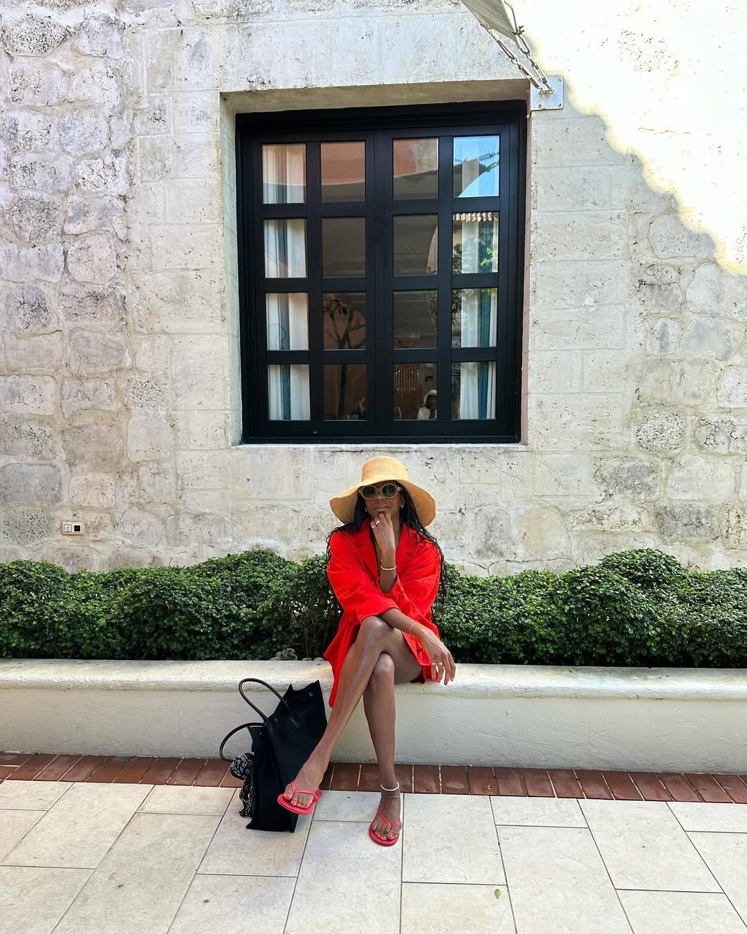 Mulher sentada com blazer vermelho oversized, chinelo vermelho de tiras finas, chapéu de palha e óculos escuros.
