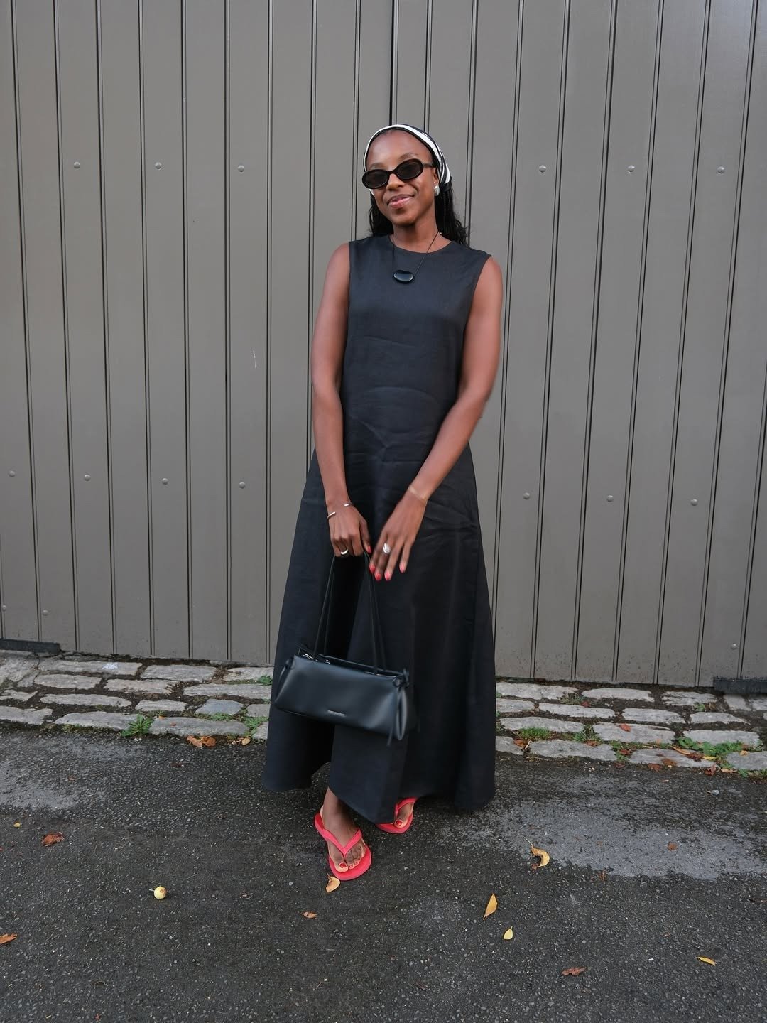 Mulher posa com vestido longo preto de modelagem reta, chinelo vermelho vibrante emborrachado e acessórios minimalistas.