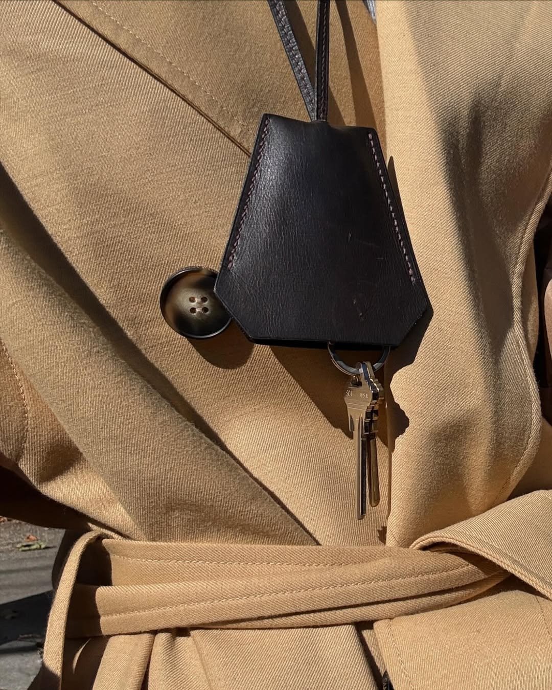 Colar funcional em couro preto com chaveiro prateado destaca-se sobre casaco bege de modelagem estruturada e cinto ajustado,