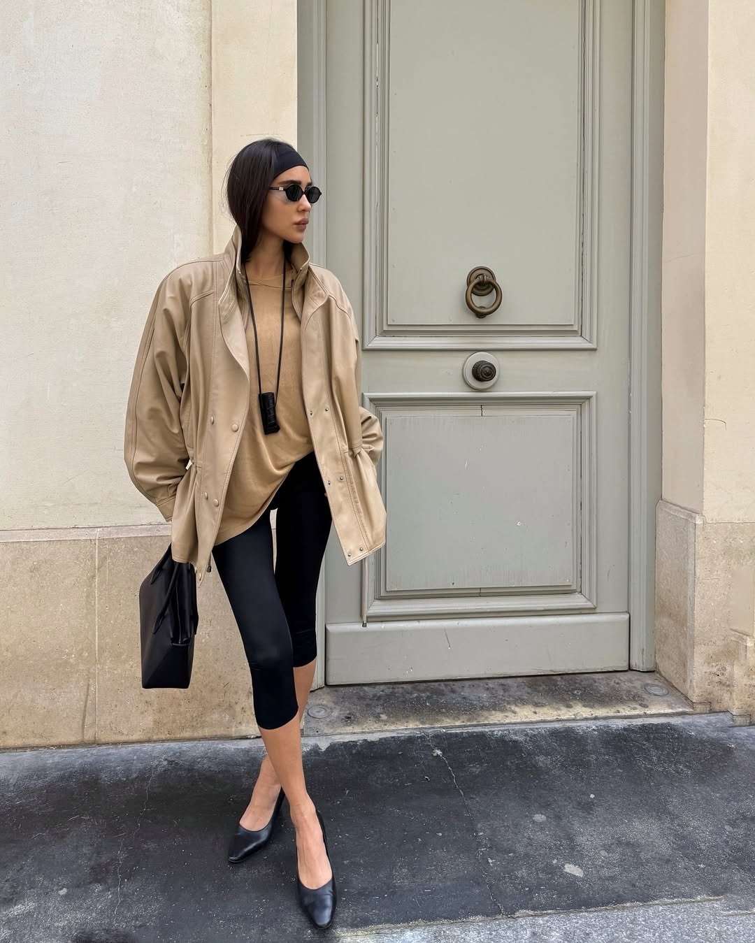 Modelo posa com colar funcional preto em cordão longo, jaqueta oversized bege de couro, legging preta e scarpin, em estilo