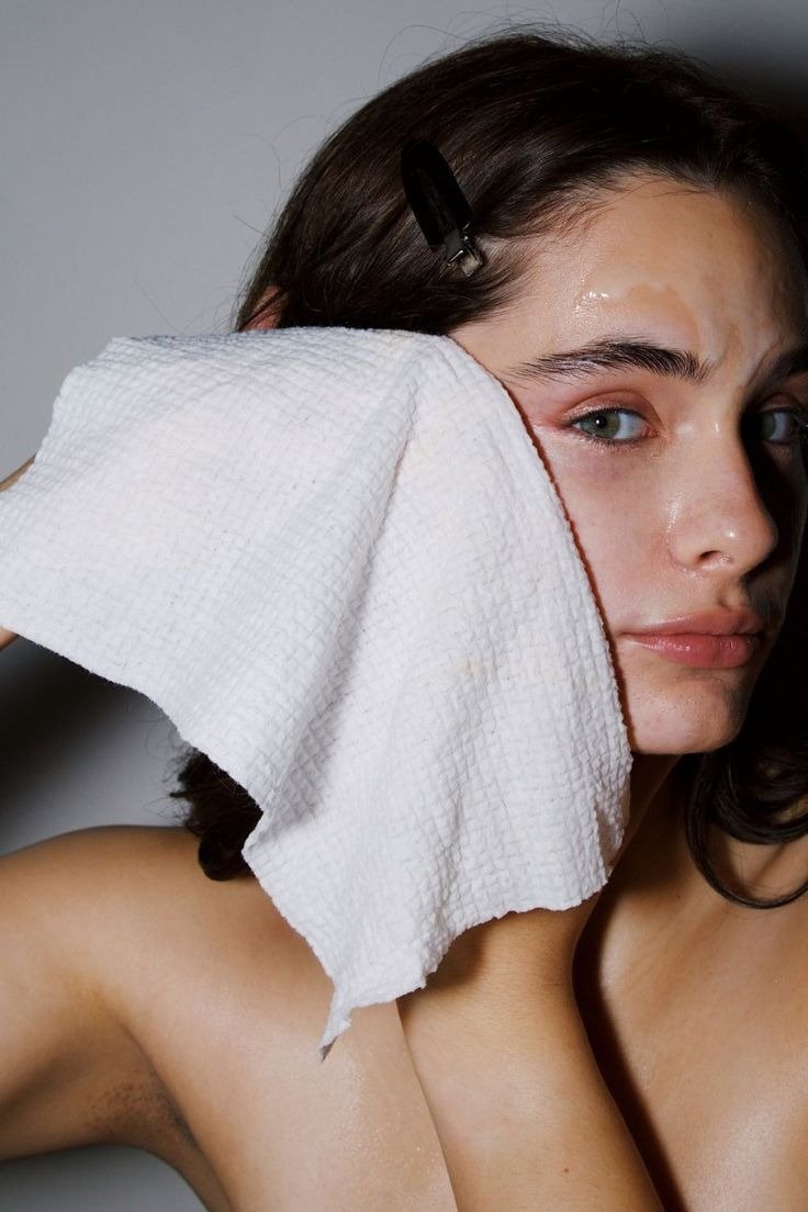 Modelo segura toalha branca texturizada no rosto úmido, demonstrando cuidado e delicadeza no processo de como limpar a pele.