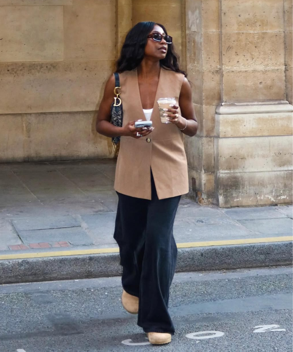 Mulher caminha confiante usando clog confortável bege, colete marrom sem mangas e calça preta ampla em cenário urbano.