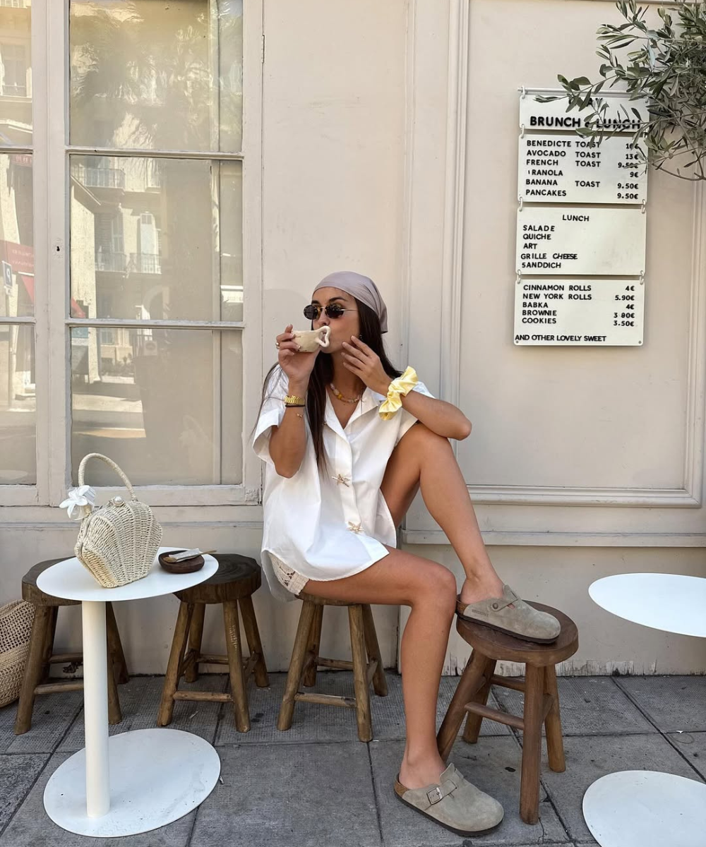 Modelo senta casualmente em banquinho de madeira com camisa branca ampla, lenço lilás na cabeça e clog confortável bege em