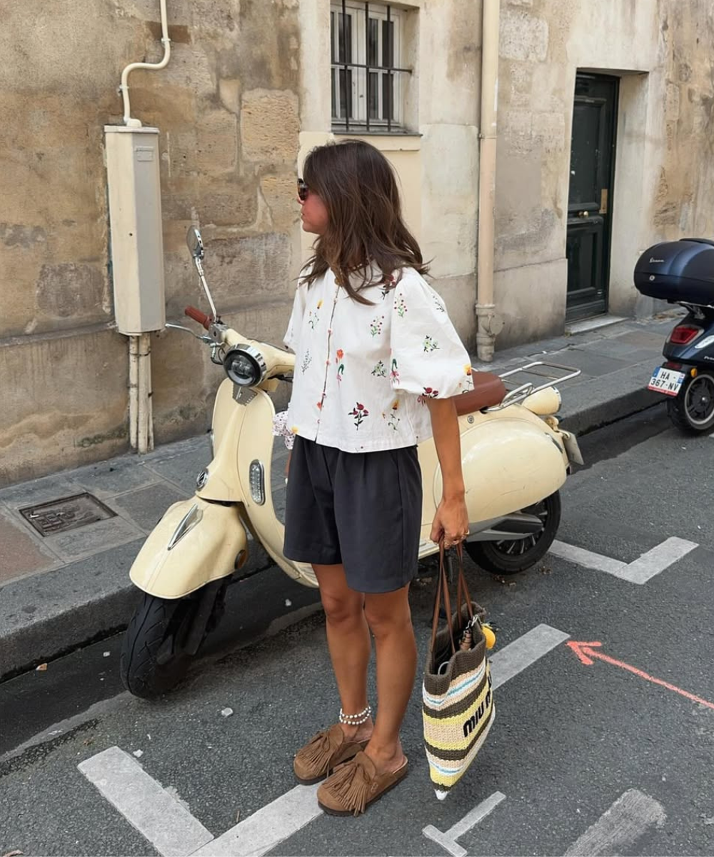 Mulher posa ao lado de scooter usando clog confortável marrom em camurça com franjas, shorts soltos e blusa branca bordada,