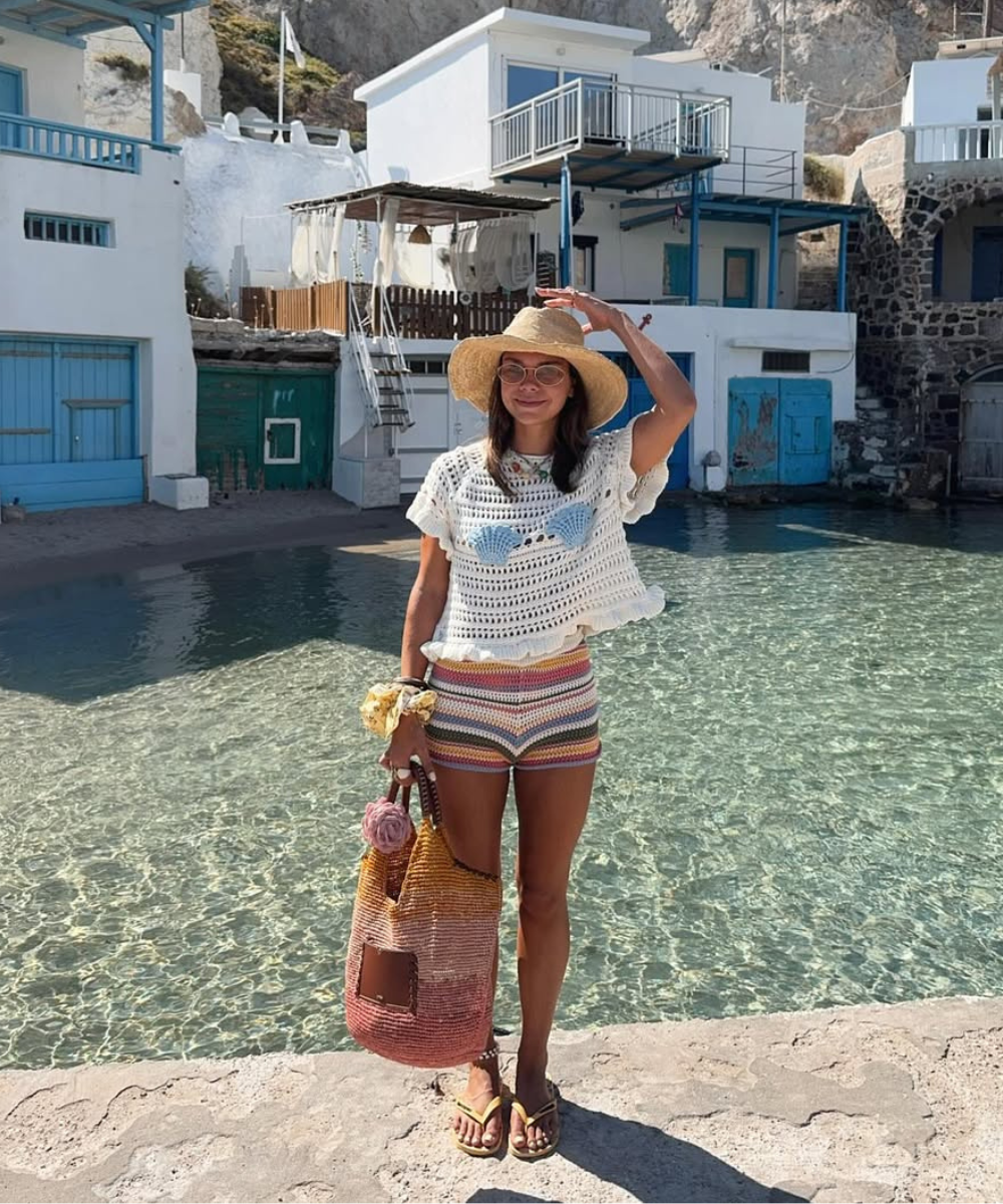 Modelo posa à beira da água usando short de crochê listrado em tons terrosos, blusa branca cropped e chapéu de palha, clima